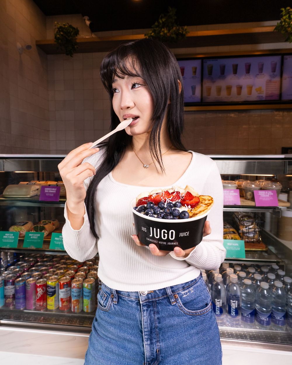 Someone in a white sweater and jeans enjoys a colorful acai bowl at a cafe with drinks display case in background.