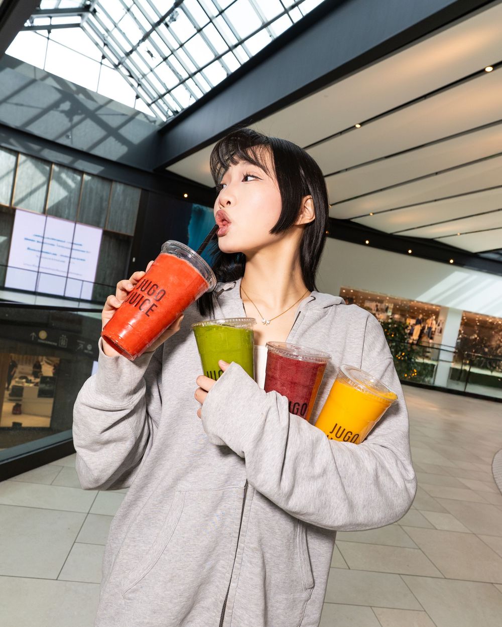Person in gray hoodie drinking colorful beverages in sequence at a modern indoor space with skylights.