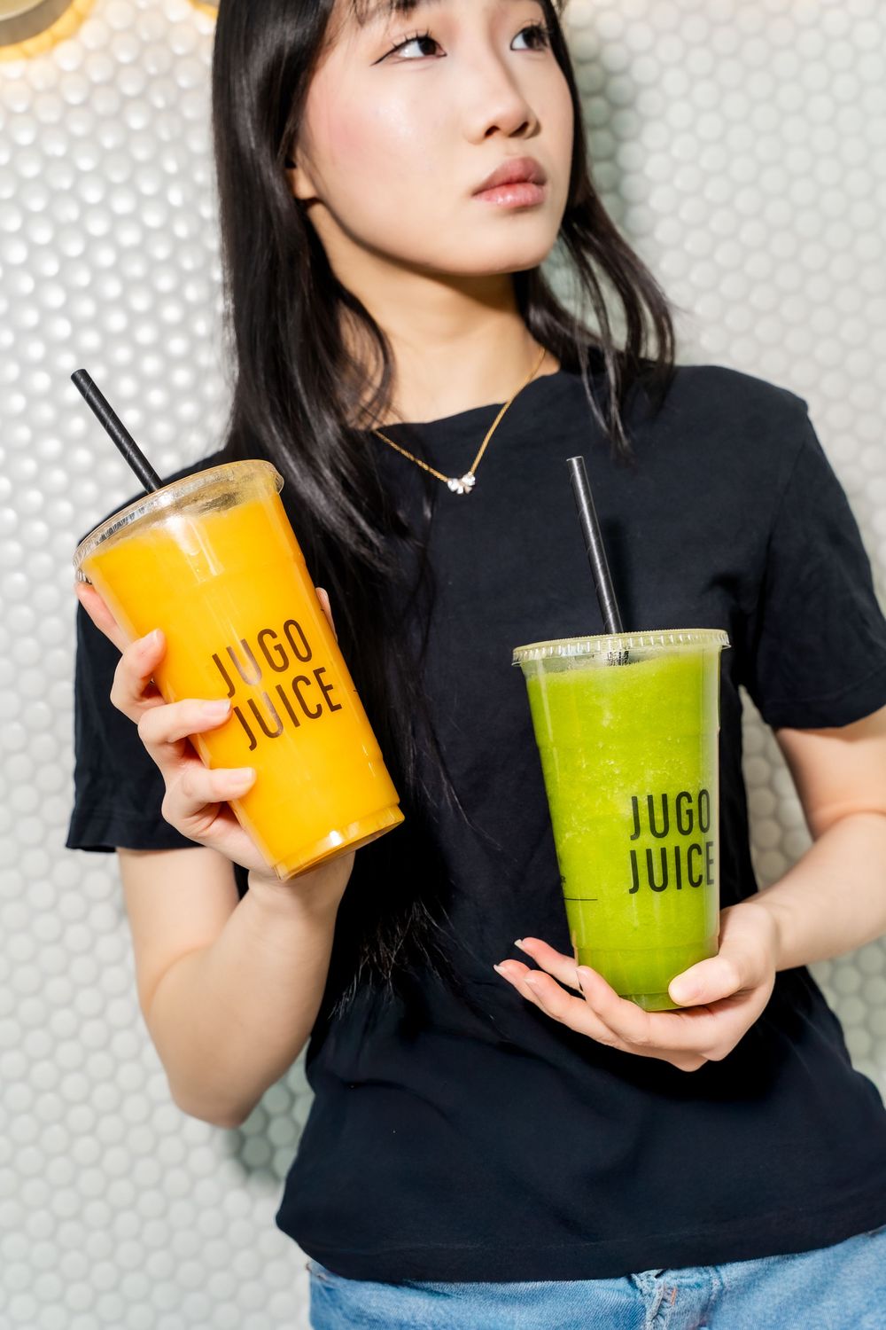 Person in black shirt holding orange and green juice drinks against white polka dot background.