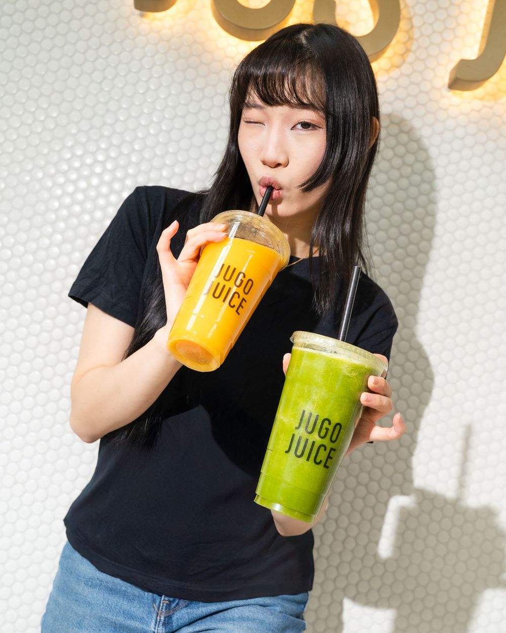 Person in black t-shirt drinking colorful beverages from plastic cups against white background.