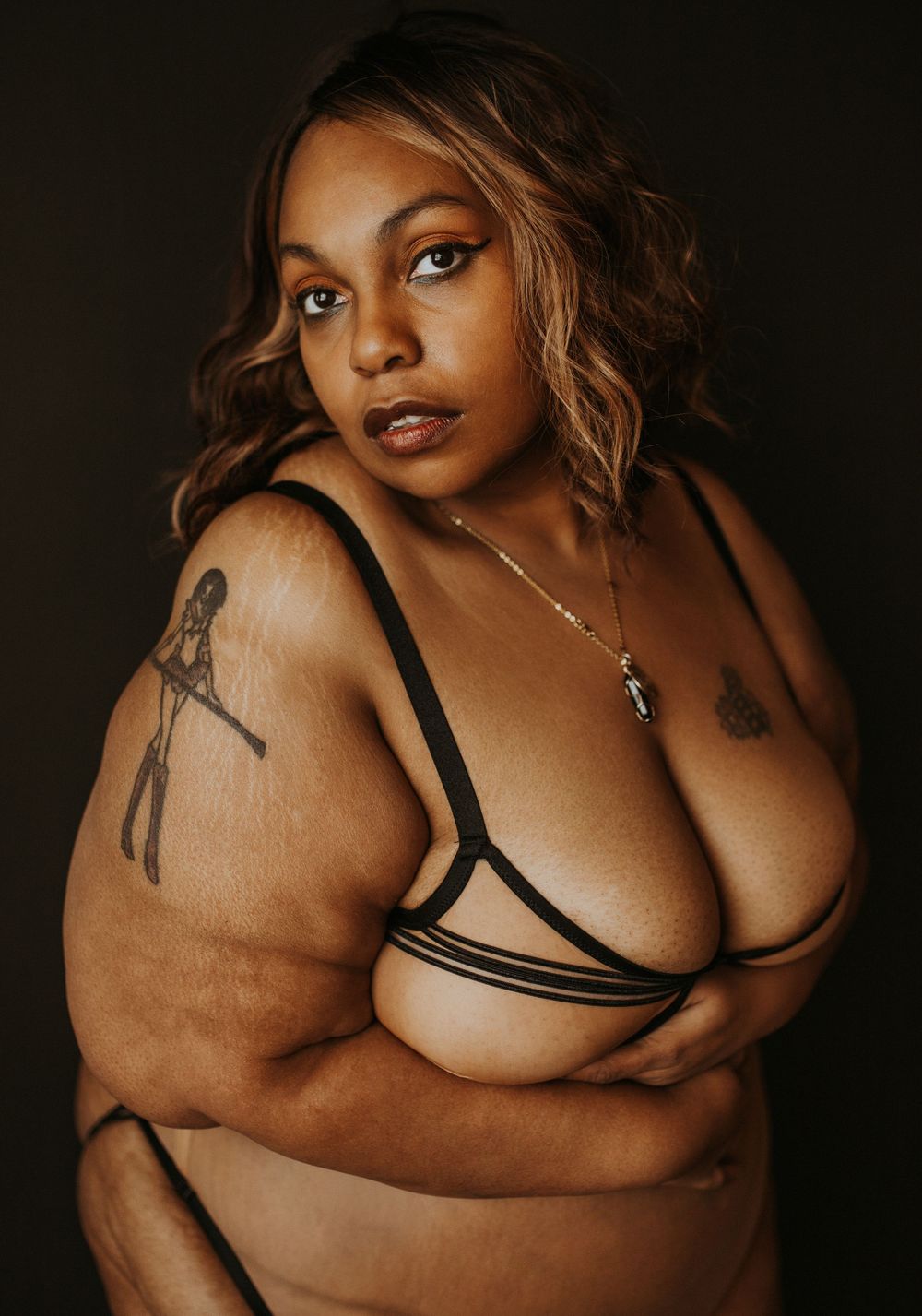 A plus-size woman poses confidently in black strappy lingerie, showcasing tattoos and natural beauty against a dark background.