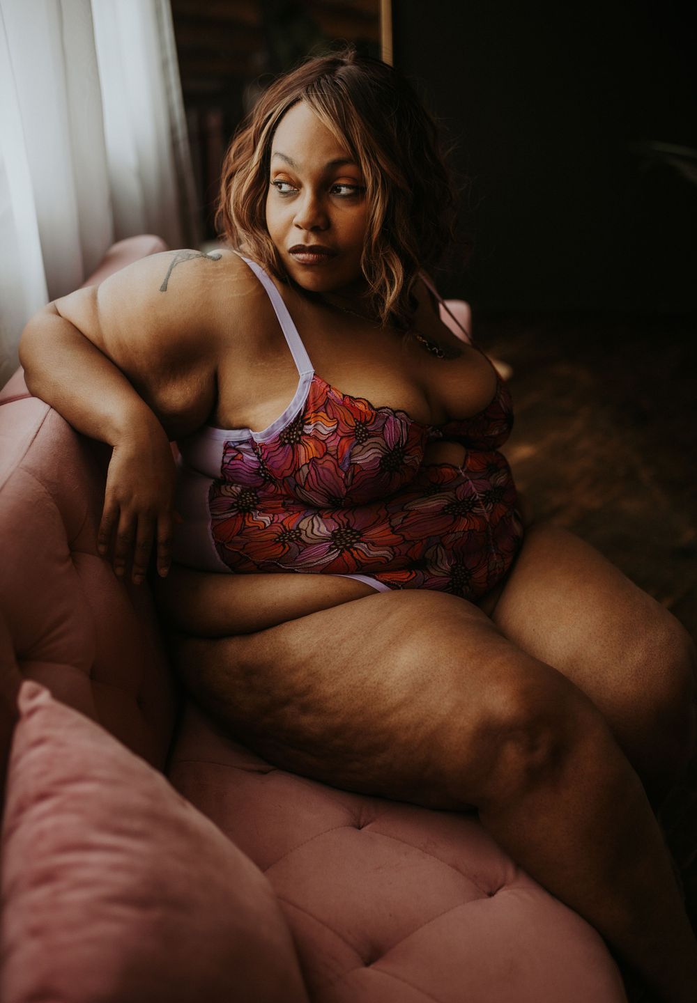 Plus-size woman in colorful floral lingerie sitting gracefully on a pink chair in a softly lit room.