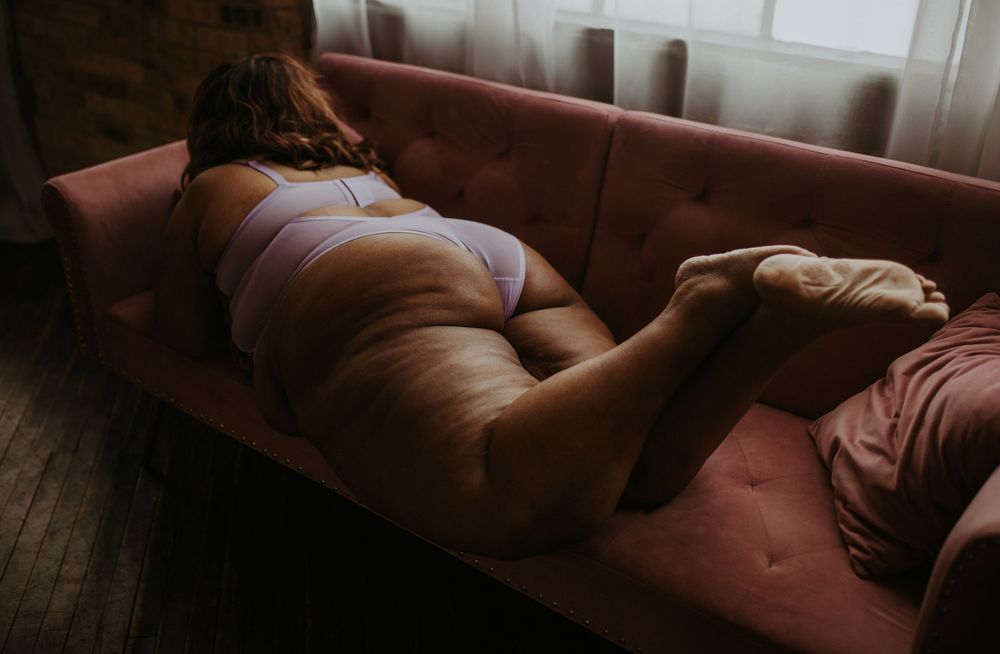 A curvy woman in lavender lingerie rests on a pink velvet sofa in a moody, dimly lit room.