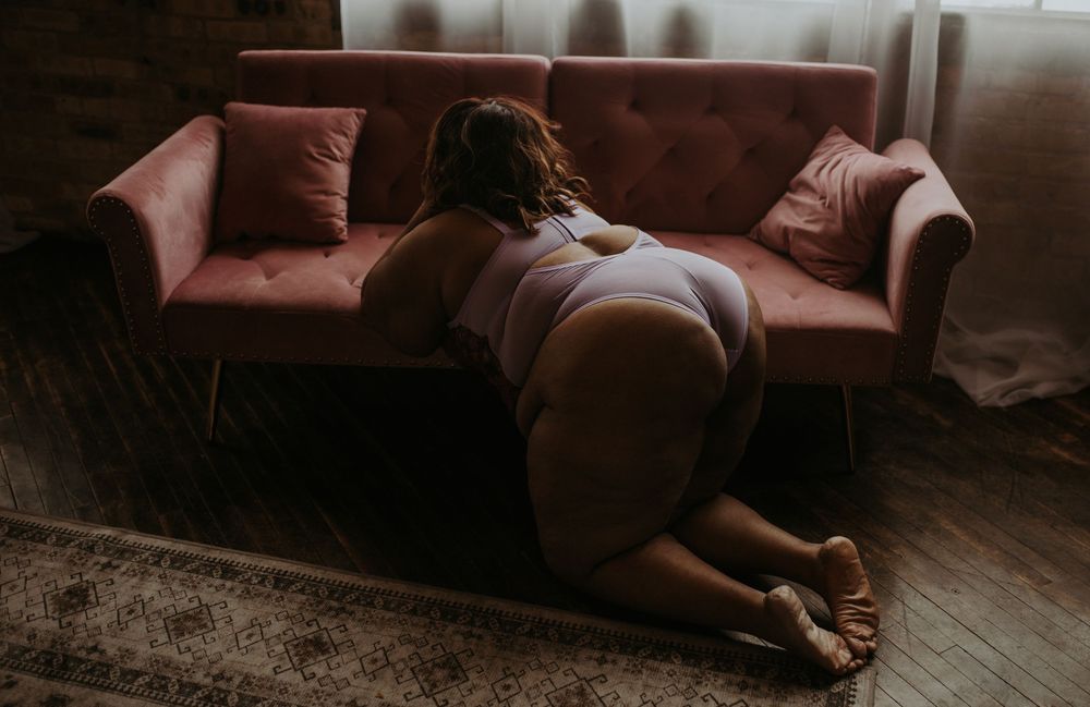 A plus-size woman in white lingerie kneeling on a wooden floor beside a pink velvet sofa.