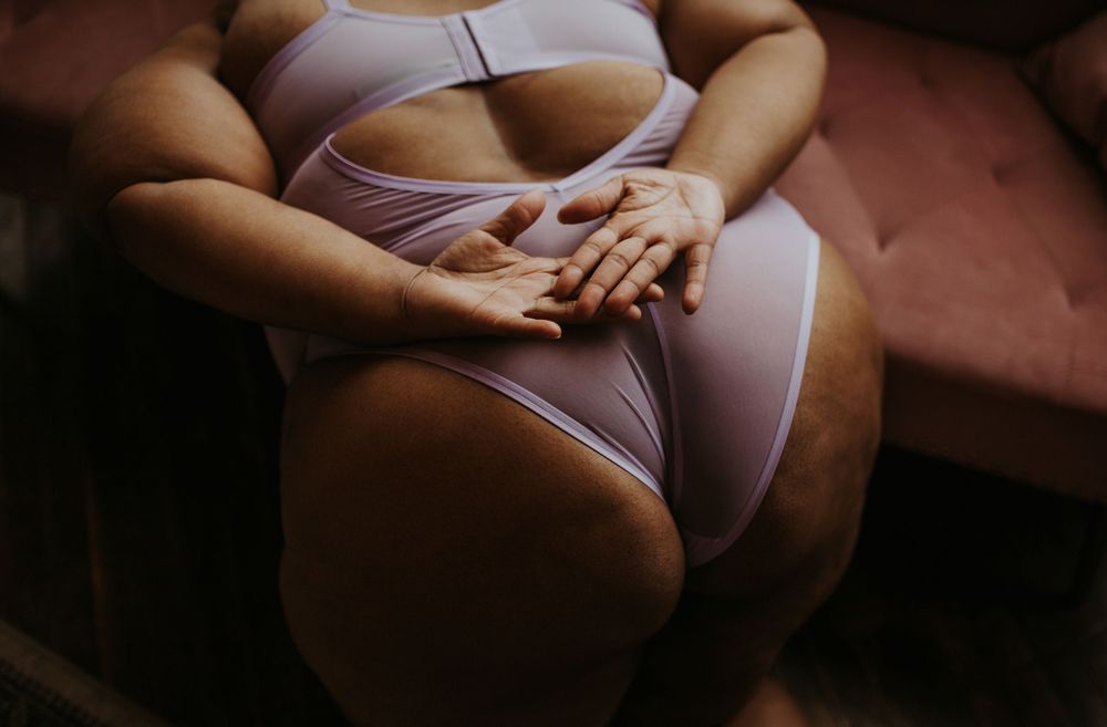 A plus-size woman in white lingerie sitting on a pink velvet sofa with hands resting on lap.