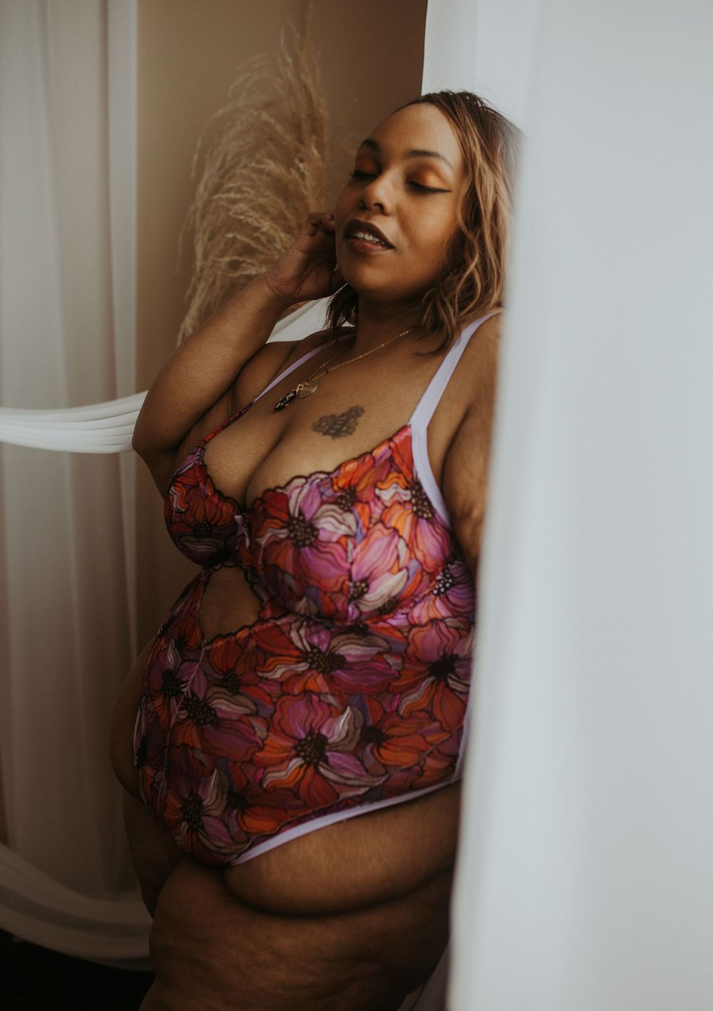 Plus-size woman in vibrant floral lingerie posing gracefully against a white wall column.