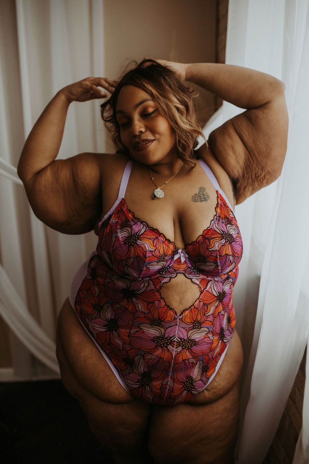 Plus-size woman in colorful floral lingerie posing confidently by a window.
