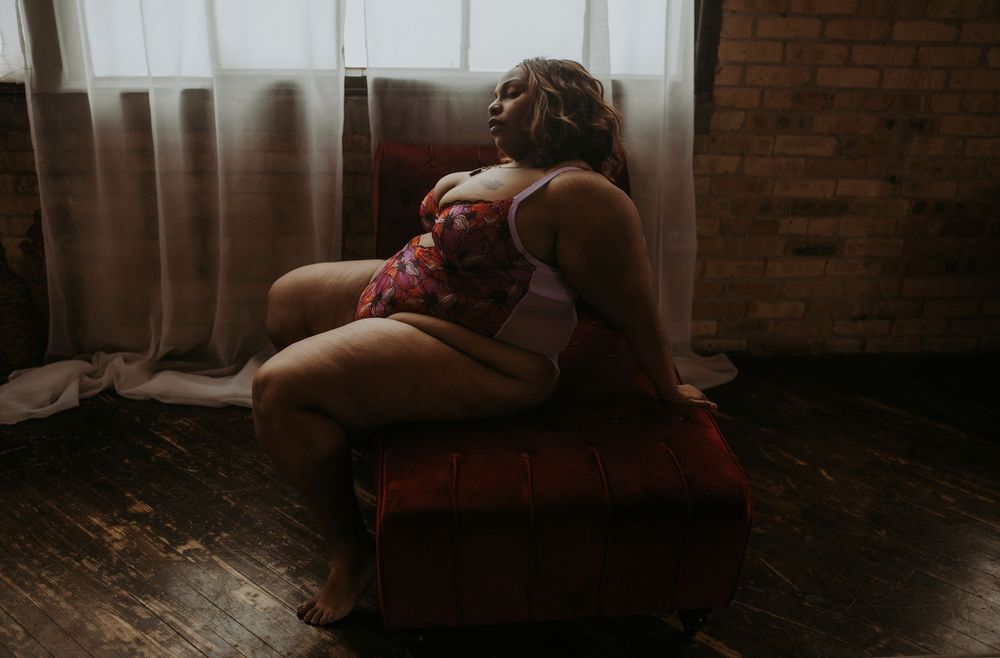 A plus-size woman poses in red floral lingerie on a vintage red velvet chaise lounge near a brick wall.