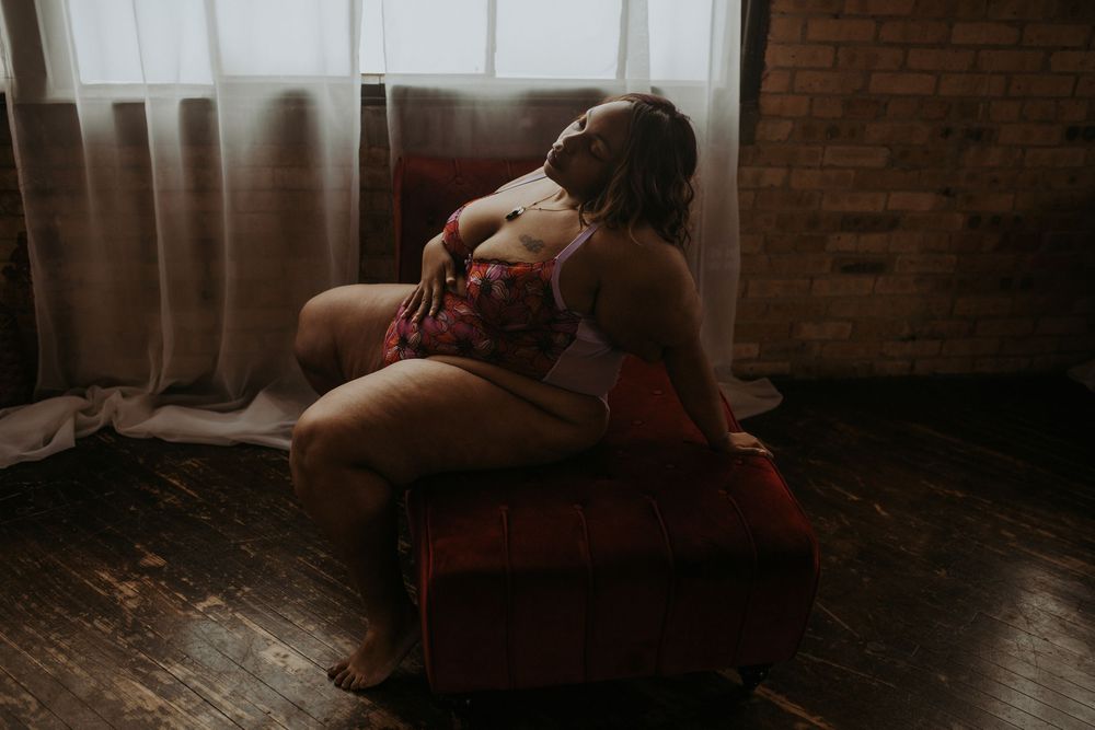 Plus-size woman in floral lingerie posing in a moody, dimly lit room with brick wall and sheer curtains.