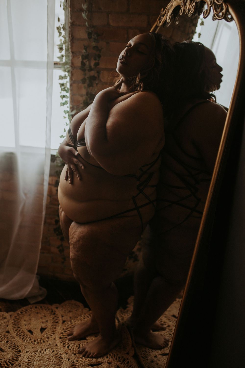 Plus-size woman in dark lingerie leaning against an ornate gold mirror near a bright window.