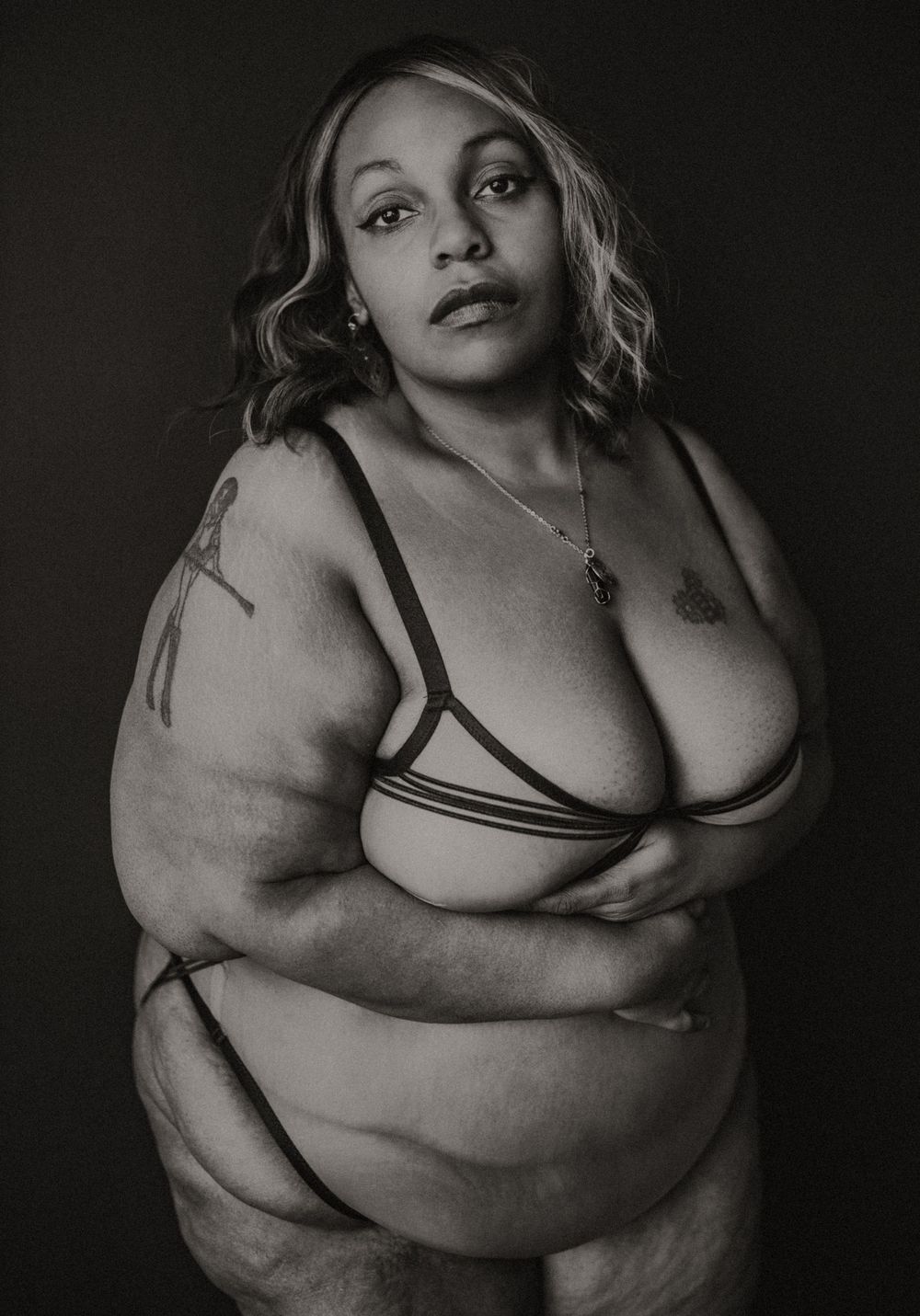 Black and white portrait of a plus-size woman in strappy lingerie posing confidently against a dark background.