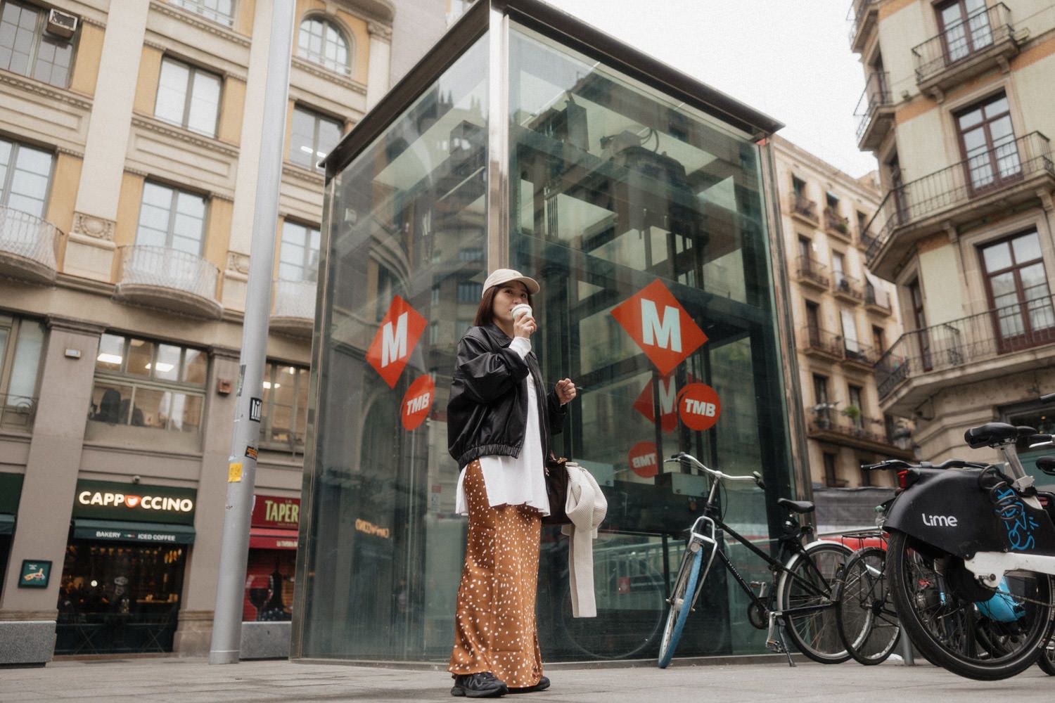 Woman in leopard print pants sipping coffee near Barcelona Metro entrance with bicycles and city buildings.