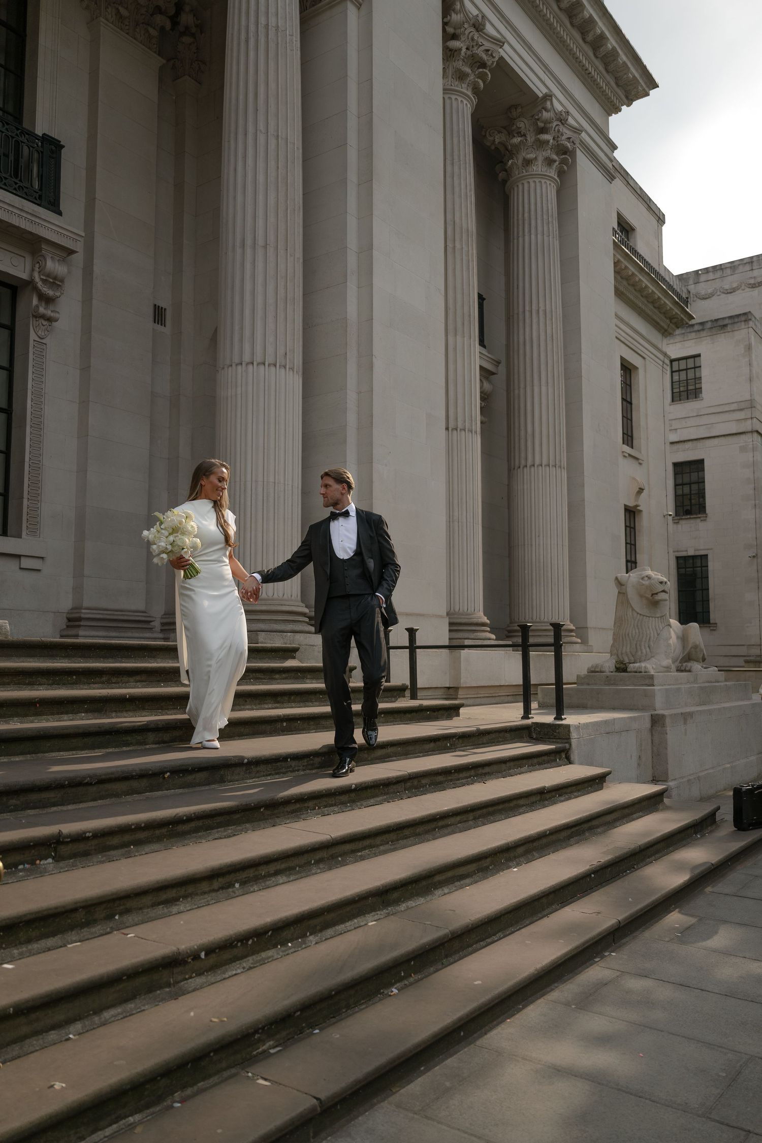 A couple descends grand stone steps outside a neoclassical building with tall white columns on their wedding day.