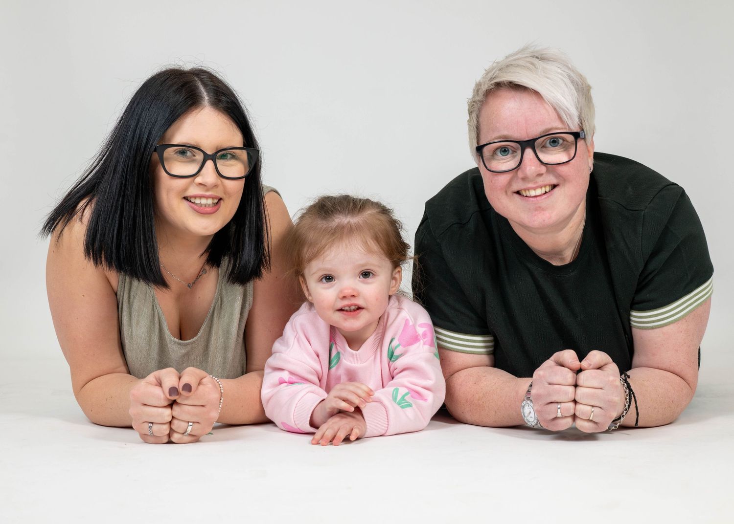 Three people wearing glasses lie on their stomachs on a white background, with a baby in pink in the middle.