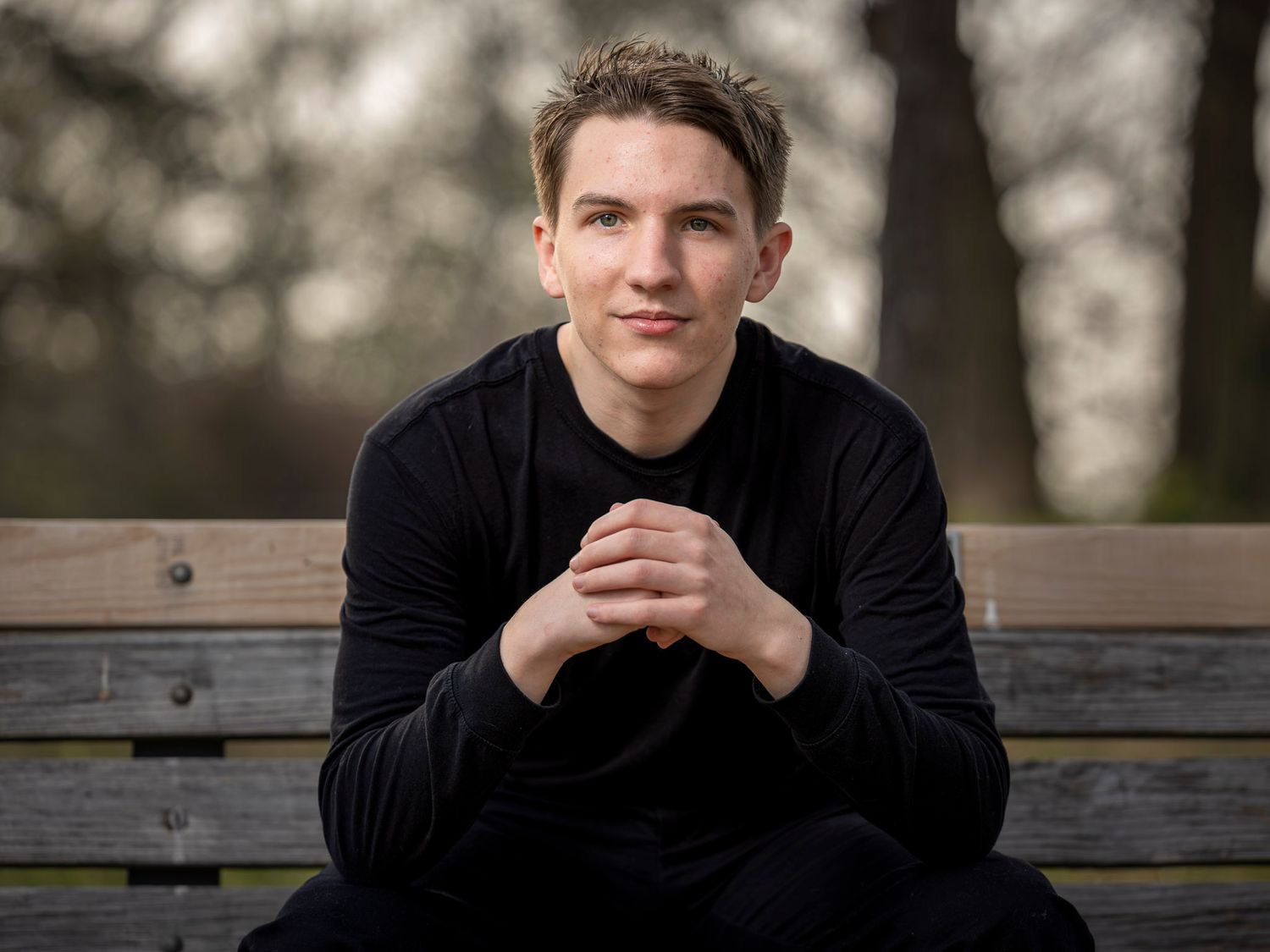 Young man in black sweater sits on a park bench with hands clasped, smiling in an outdoor setting.