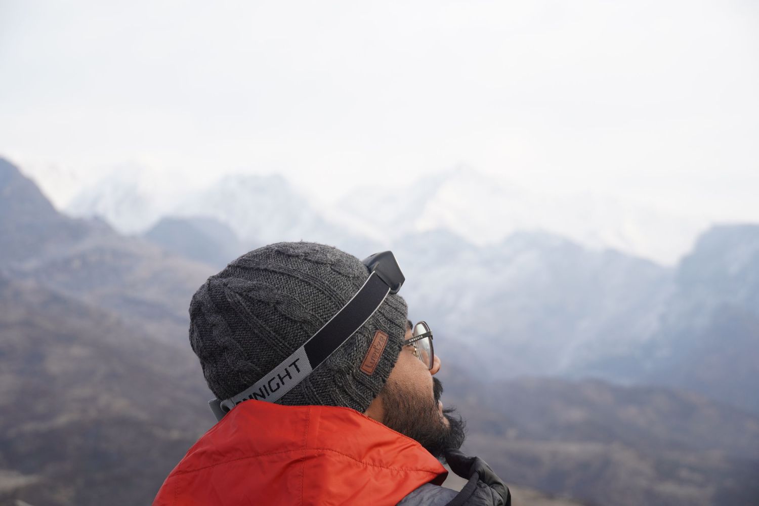 Close-up of a trekker in a red jacket looking out at snow-capped mountain peaks, a moment of witness and reflection.