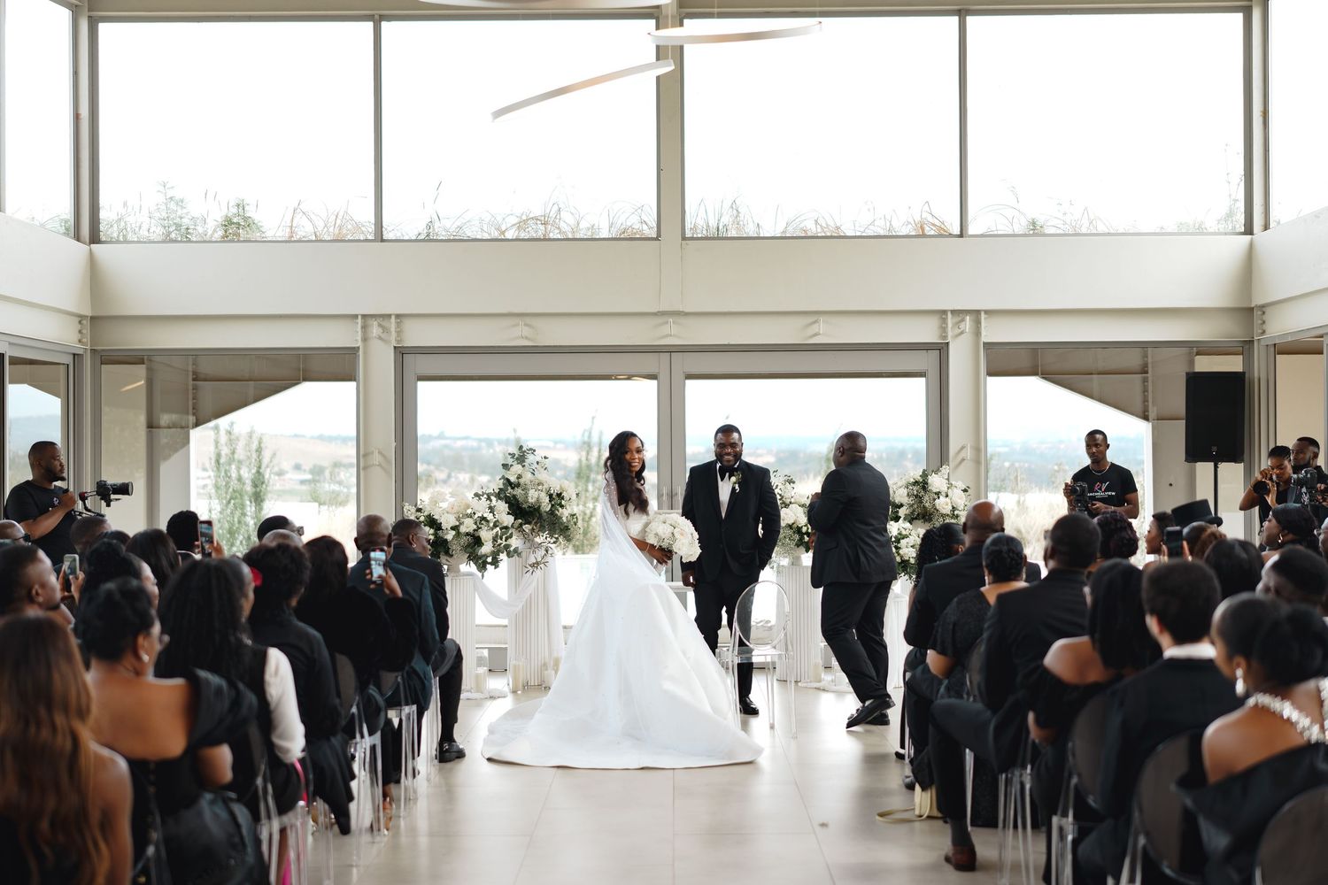 Elegant indoor wedding ceremony with bride and groom at the altar, guests seated in rows, large windows with city views.