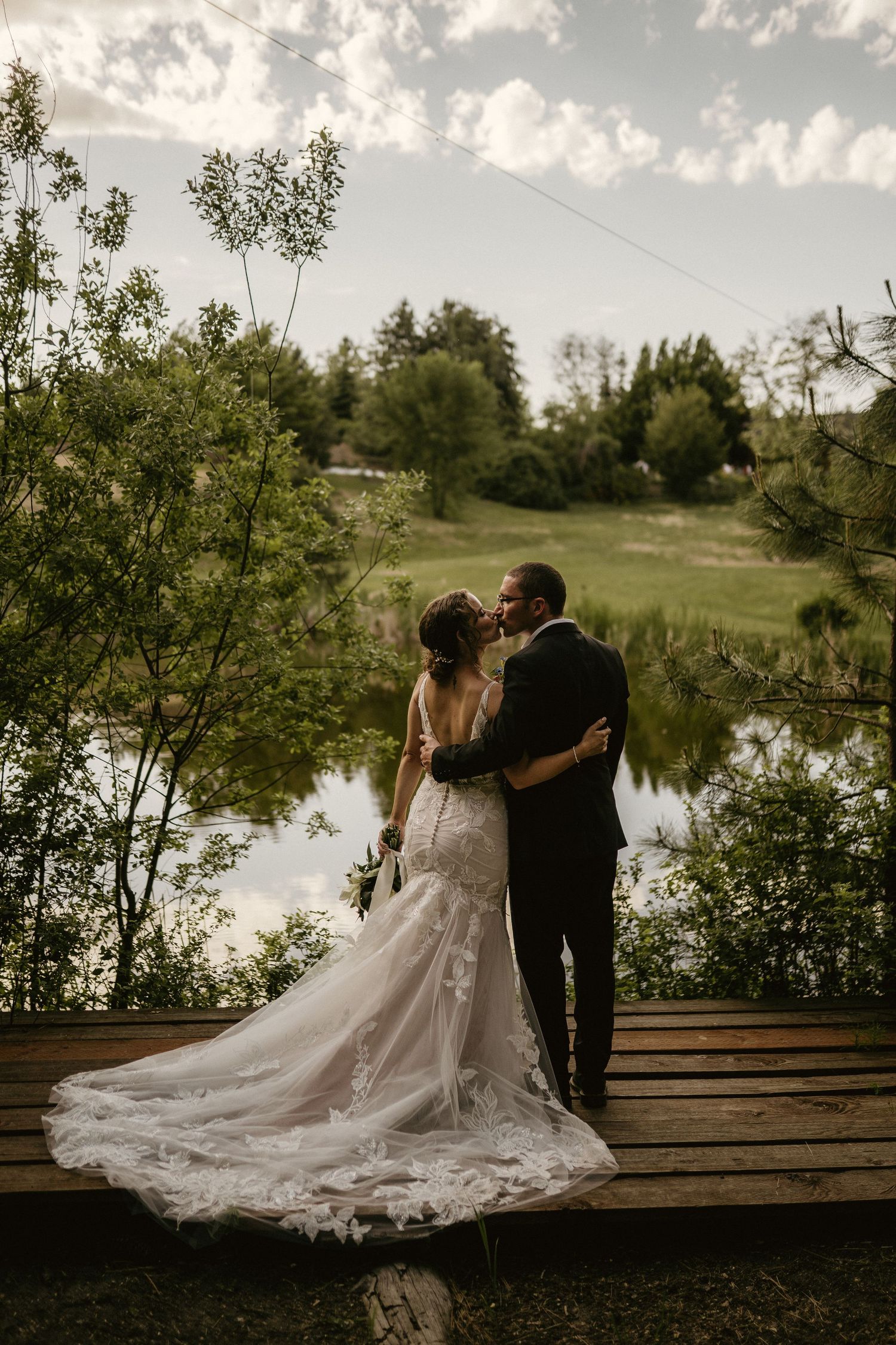 Wedding couple posing on wooden dock by lake with flowing dress train.