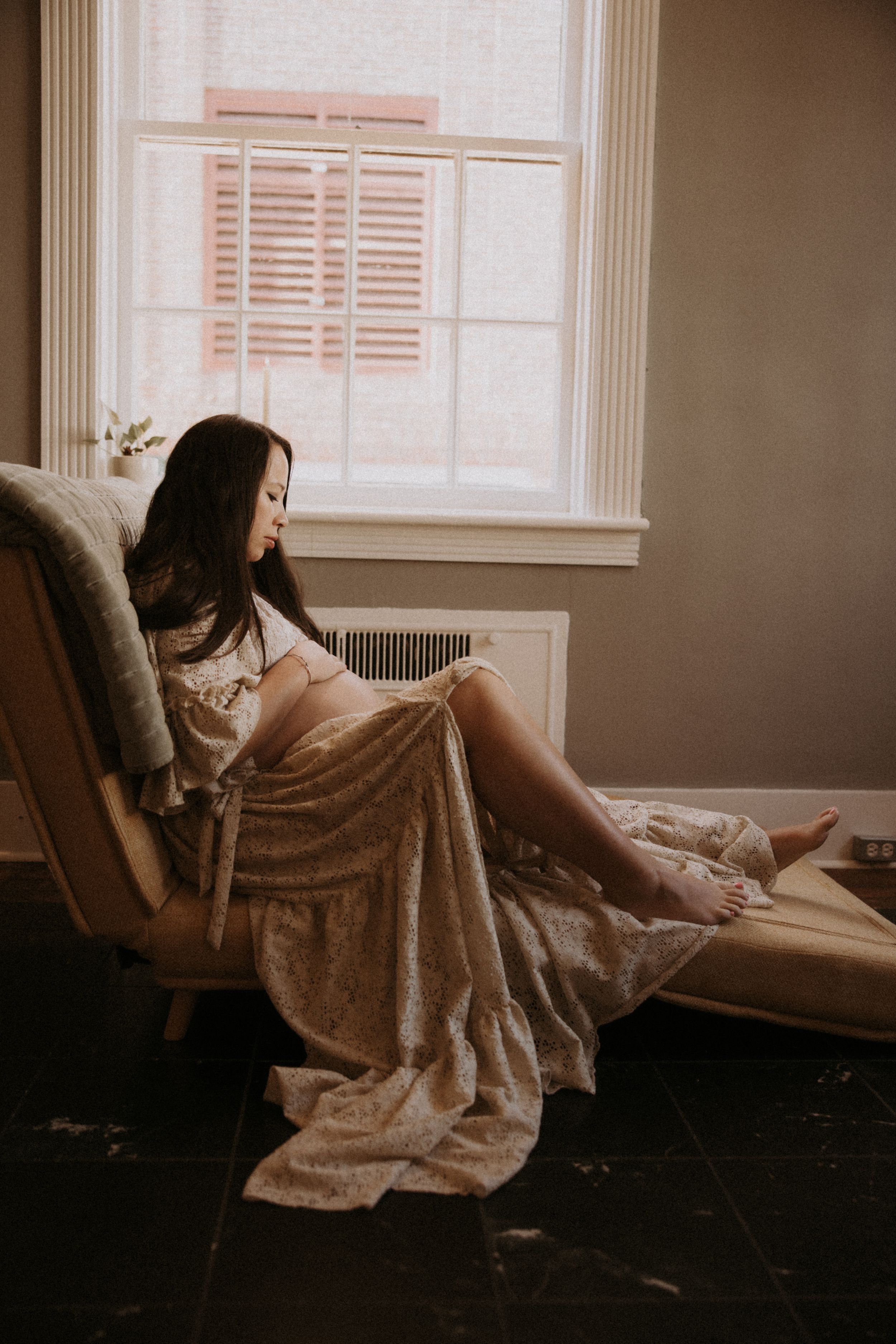 A person in a flowing dress reclines on a window seat in moody lighting creating a dreamy atmosphere.