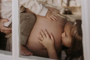 A tender moment captured through window glass as small hands touch a pregnant belly in warm, muted tones.