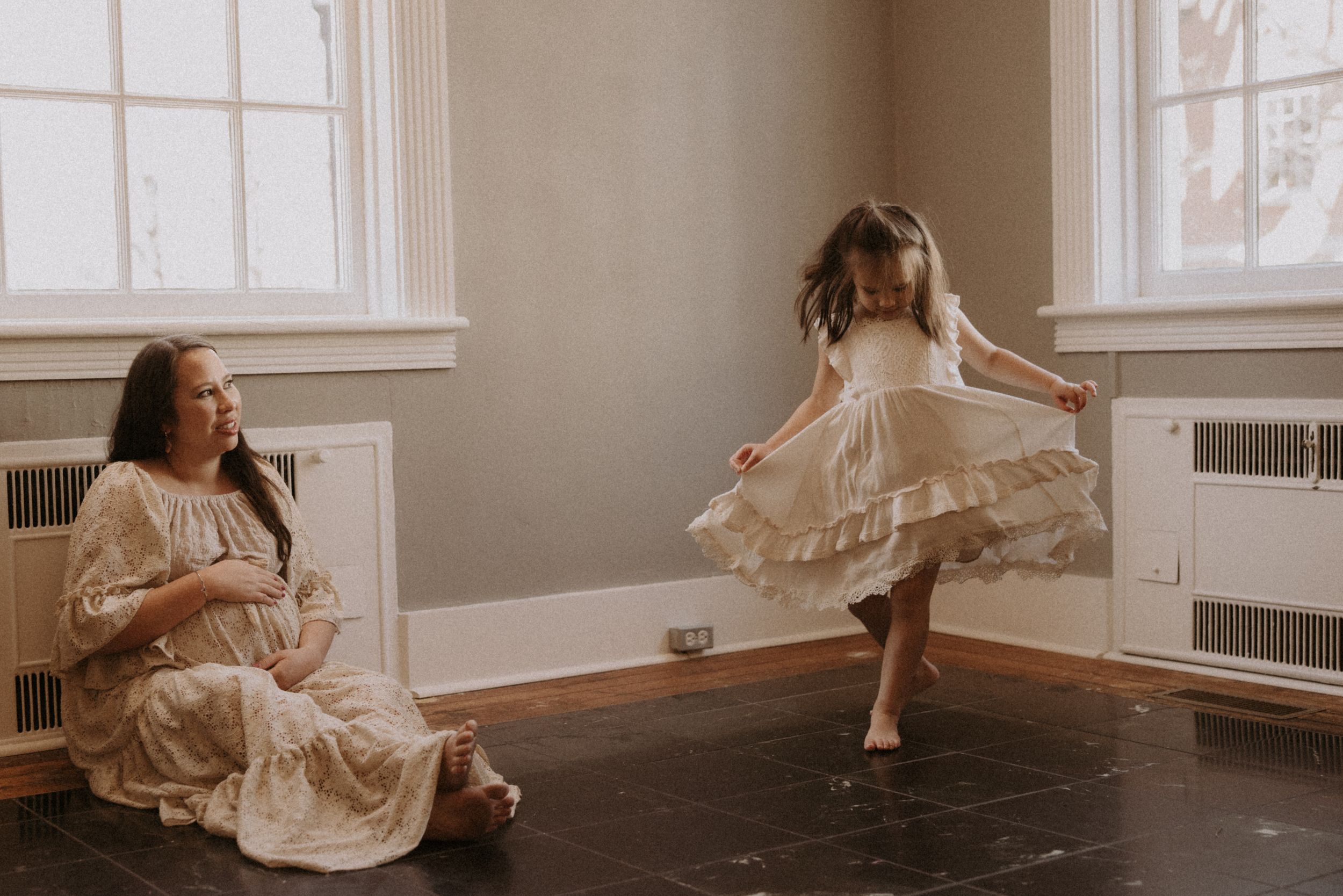 Two people in cream-colored dresses share a joyful moment dancing and twirling in a sunlit room.