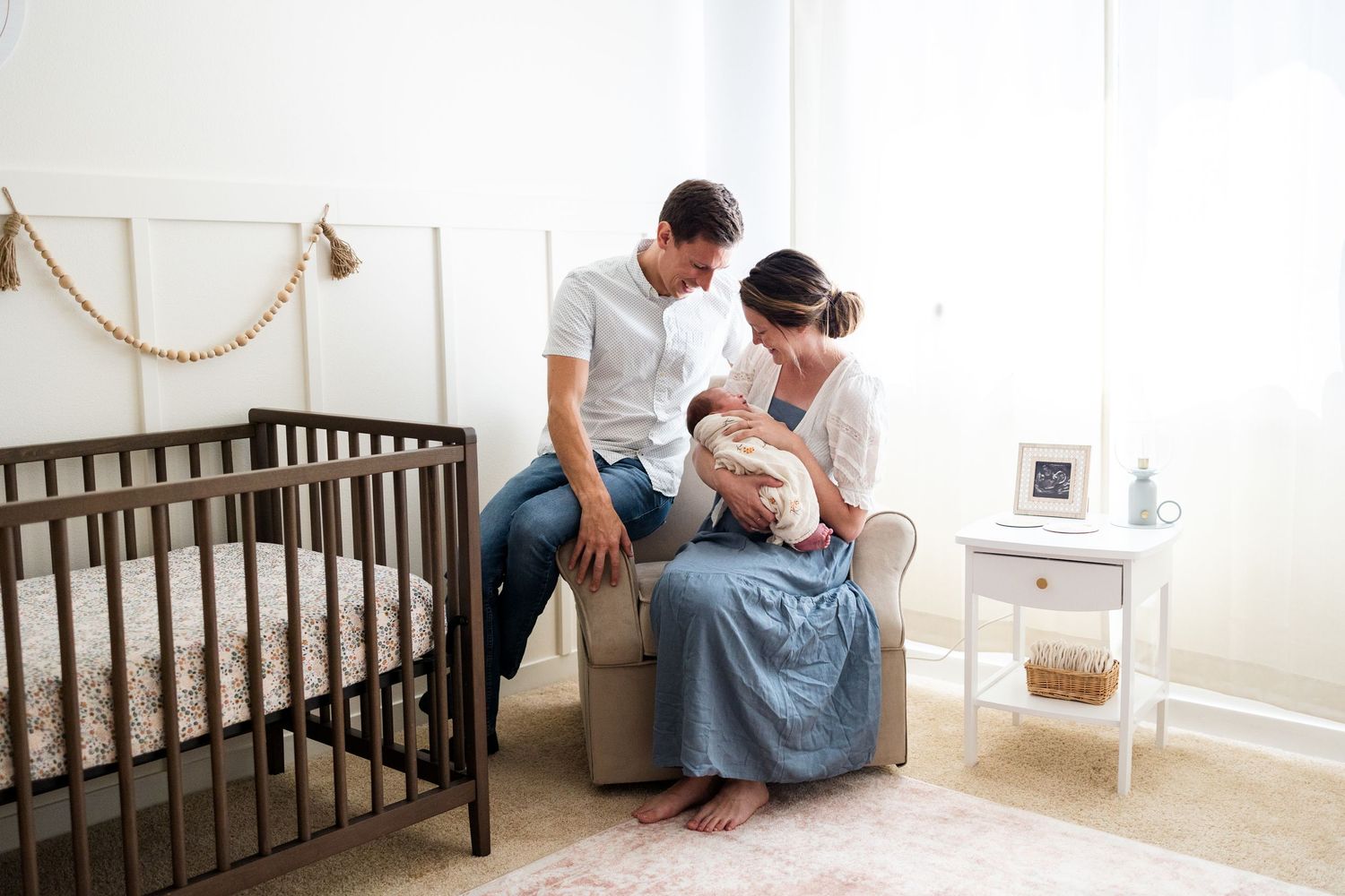 Parents sit together on a rocking chair next to a dark wooden crib in a bright nursery room.