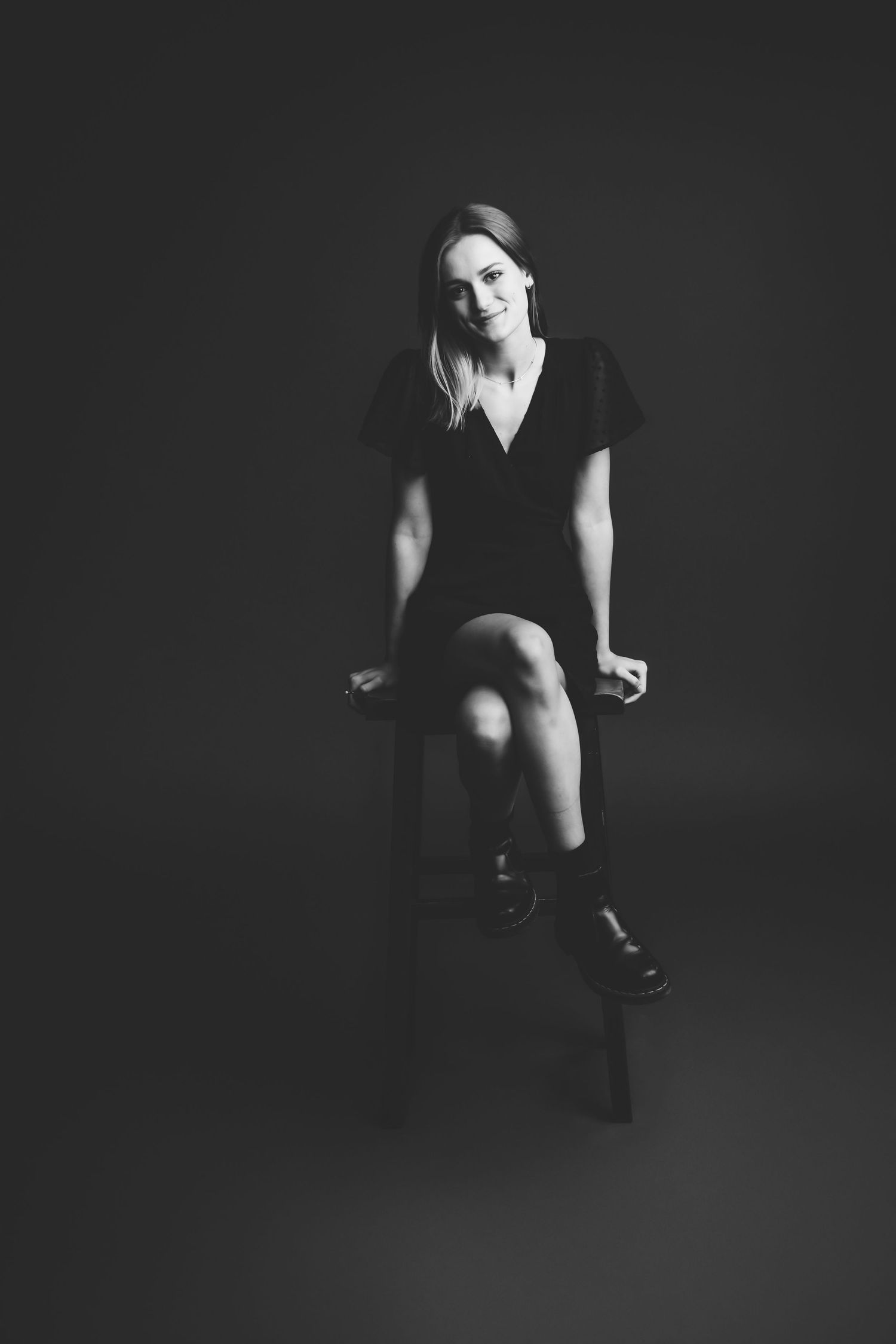 Black and white studio portrait of a woman in a black dress and boots sitting on a stool, smiling softly.