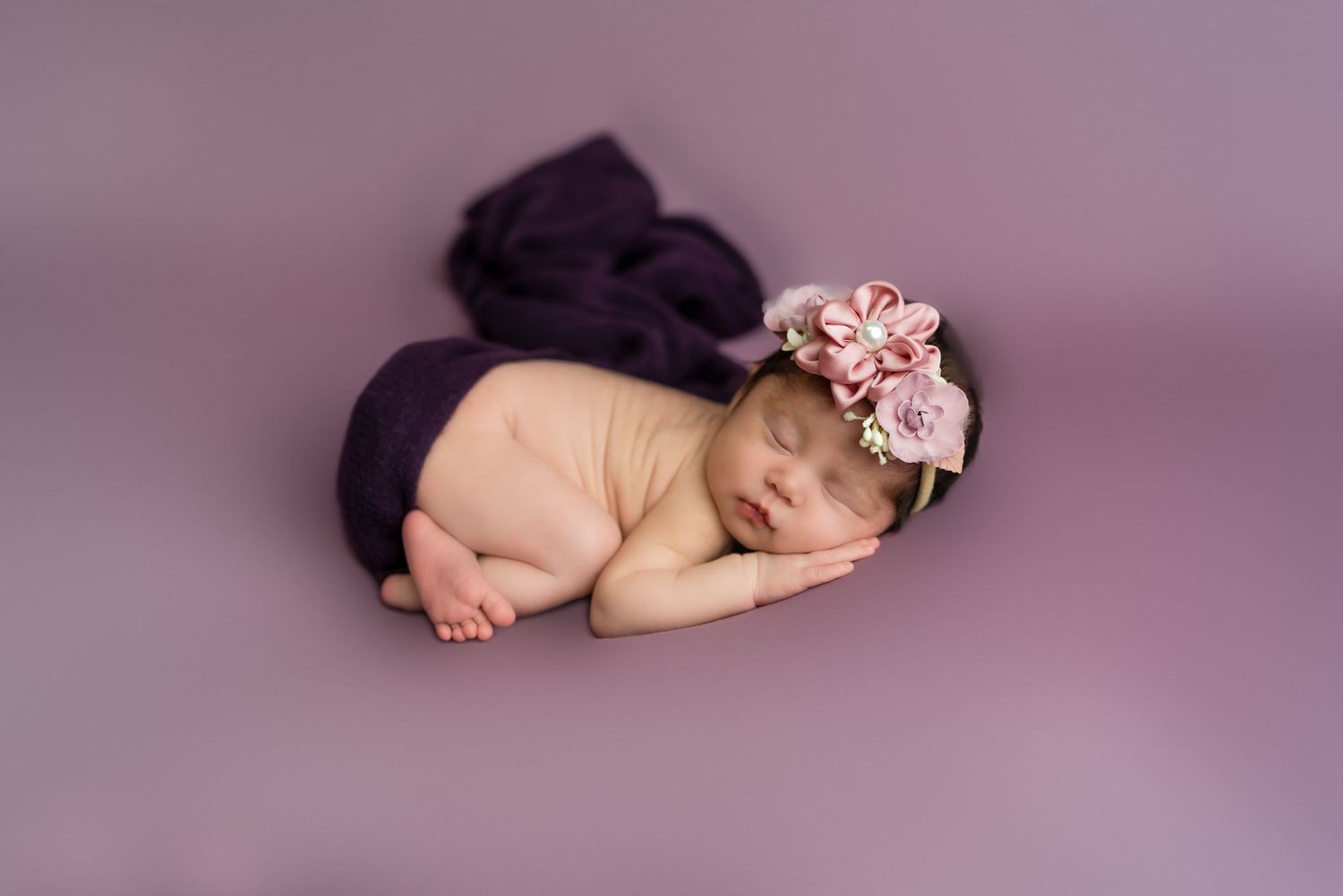 Sleeping newborn baby wrapped in soft pink accessories on purple backdrop.