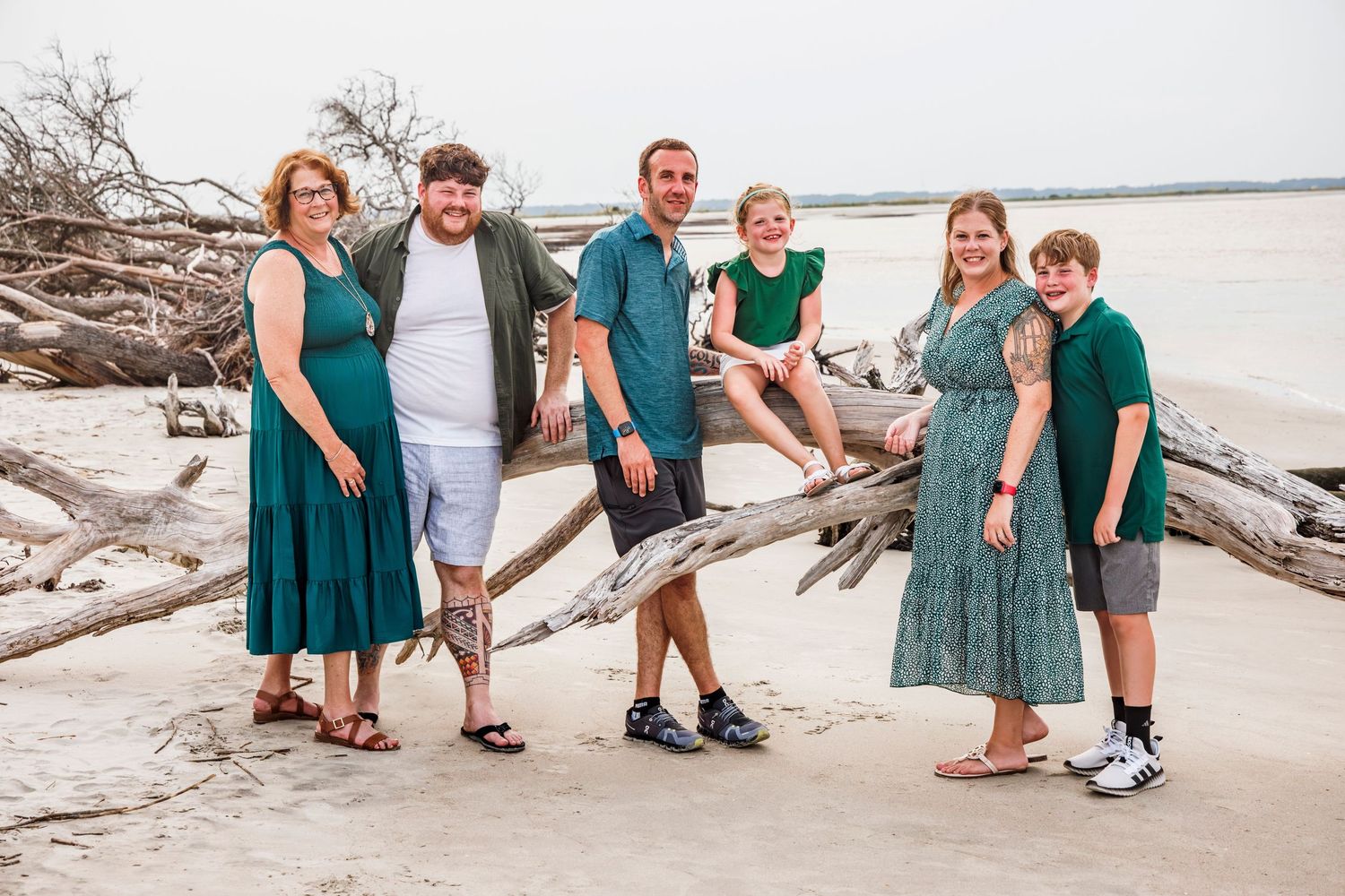A group in coordinating teal and green outfits poses together on a beach with driftwood and ocean views.