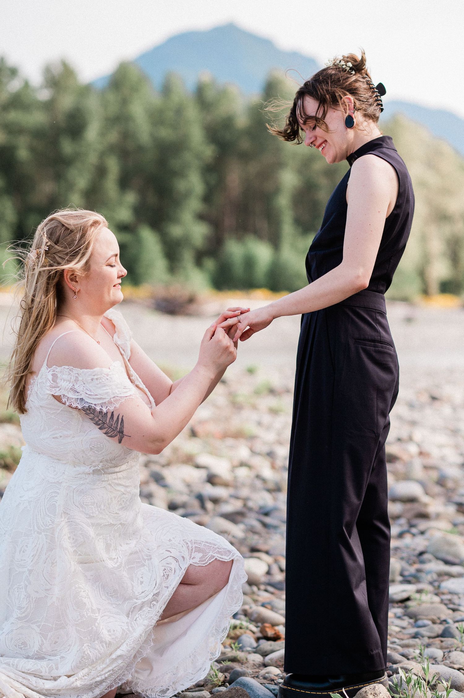 A bride goes down on one knee during elopement ceremony on a rocky riverbank with mountains in the background.