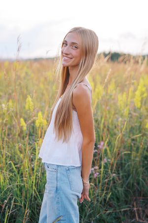 Young woman in white tank top and jeans poses in a golden field at sunset.