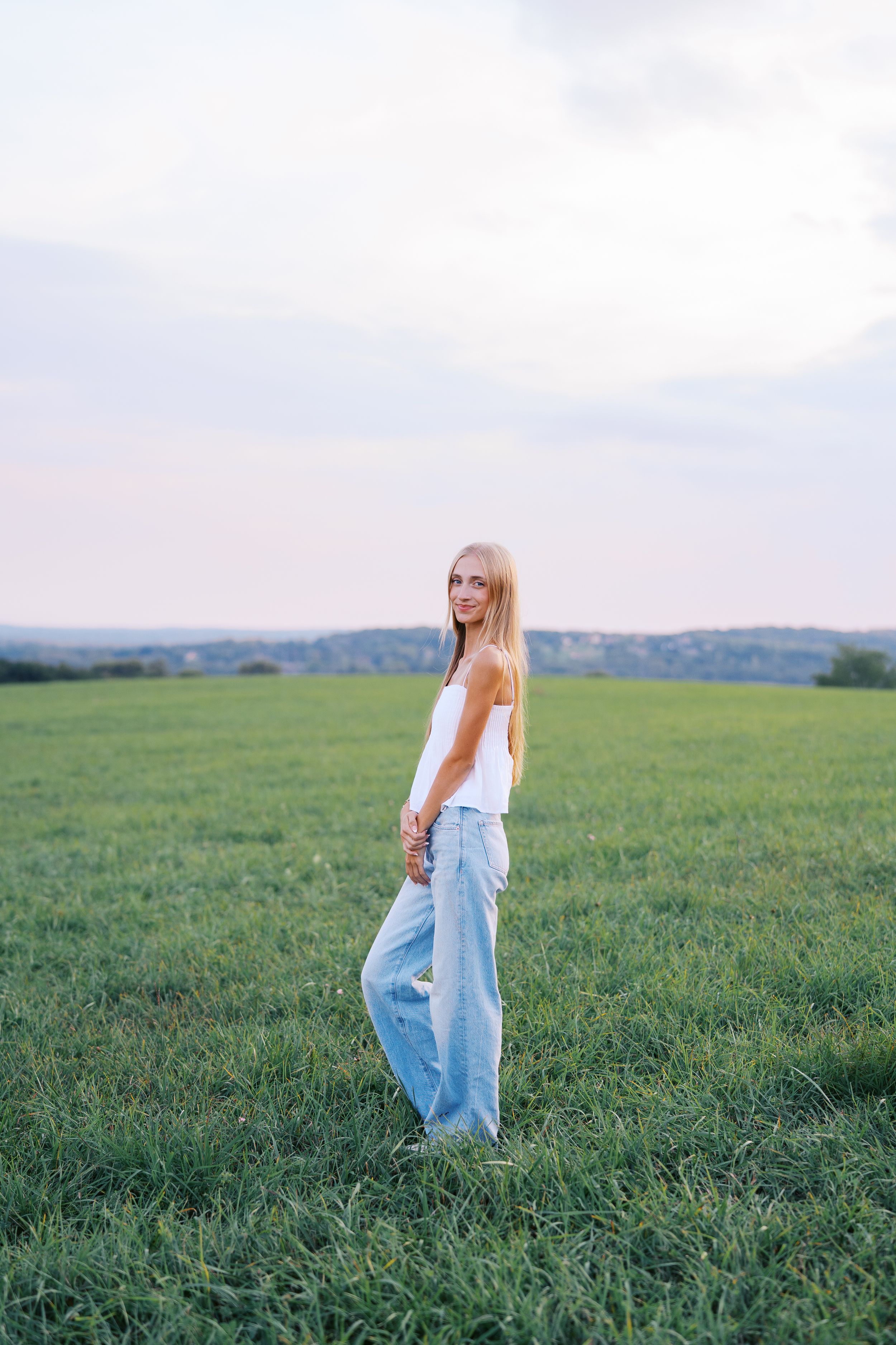 Woman in casual summer outfit stands in green field with scenic landscape.