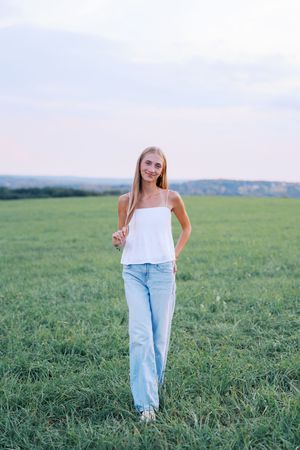 Woman in white tank top and jeans stands confidently in open field.