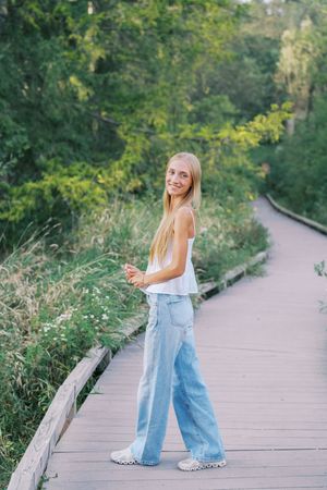 Someone in denim and white poses playfully on a wooden boardwalk surrounded by greenery.
