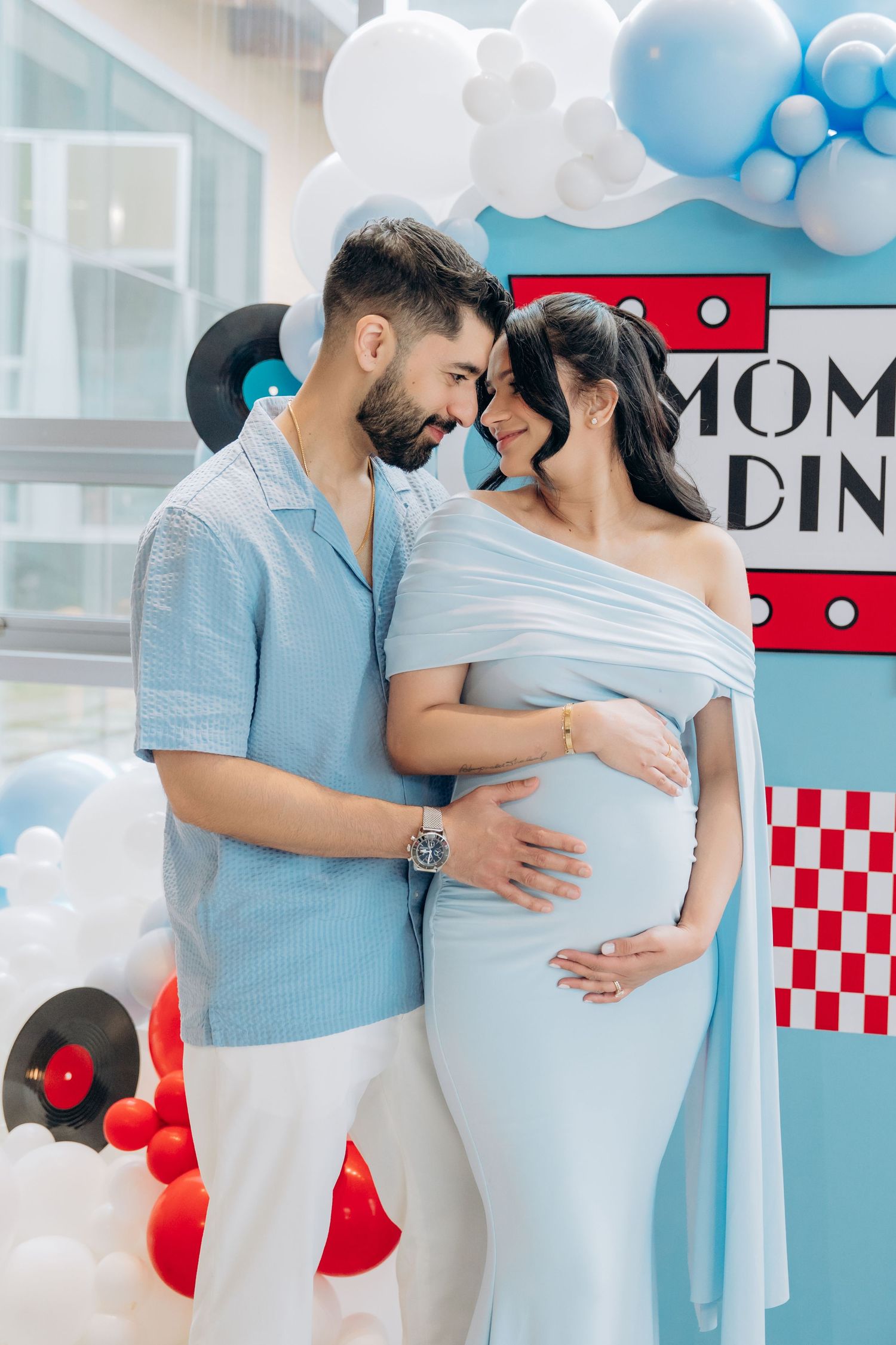 A couple in matching light blue and white outfits embrace during a pregnancy photoshoot with red and blue decor.