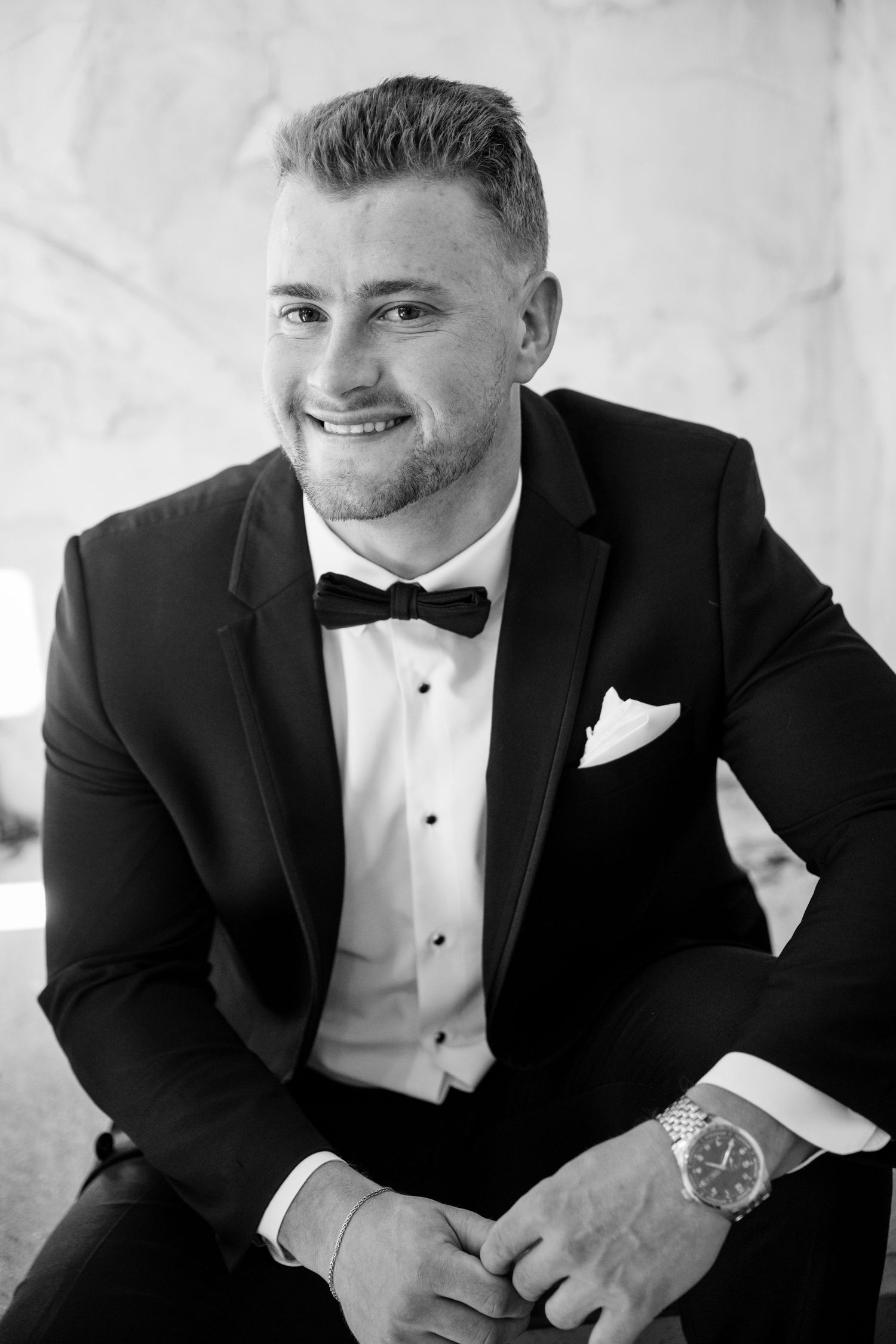 A groom in a classic black tuxedo and bowtie smiles while sitting in a candid black and white portrait series.