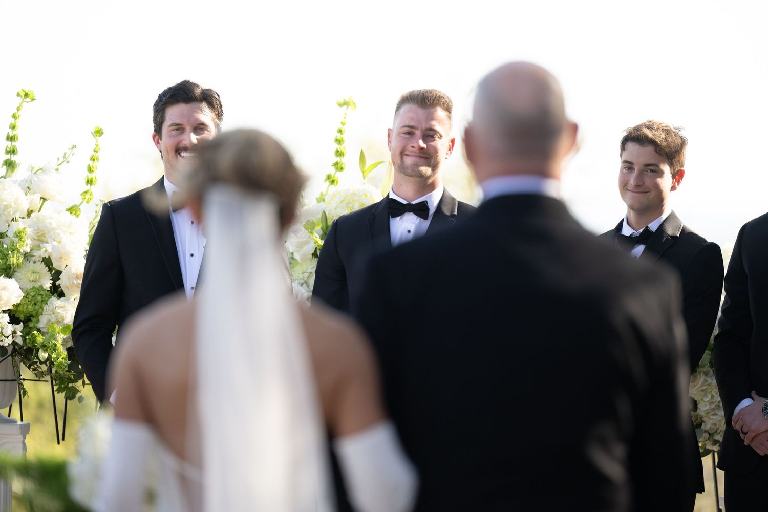 A wedding ceremony takes place outdoors with guests gathered around a couple exchanging vows in a garden setting.