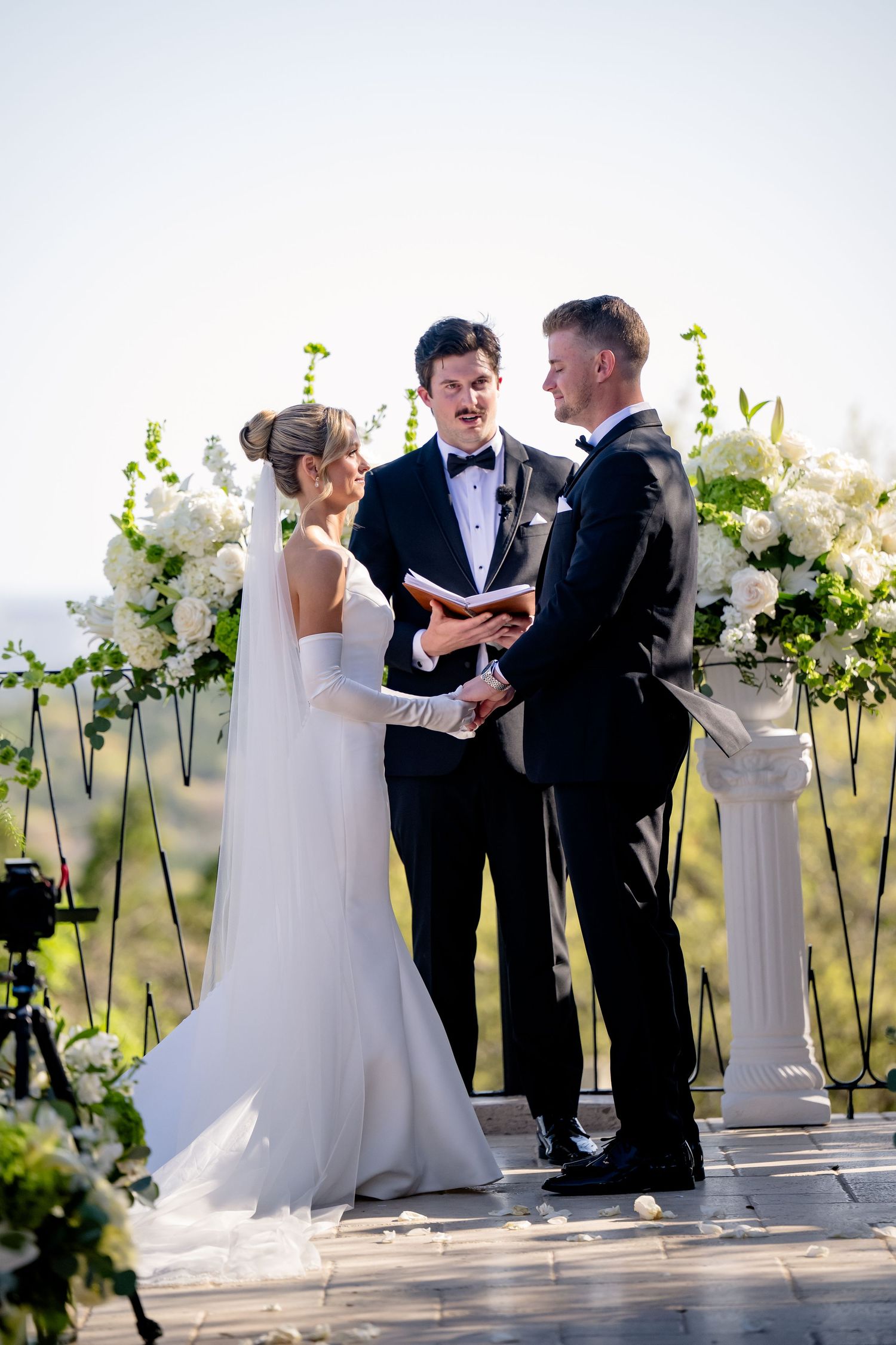 Elegant outdoor wedding ceremony with white floral arrangements on pillars and a couple exchanging vows at sunset.