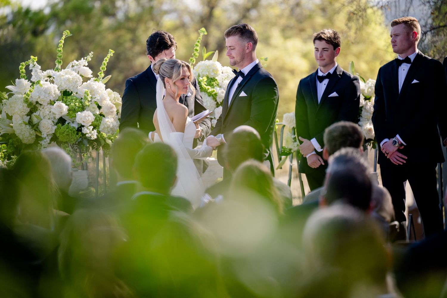 Wedding ceremony guests gather outdoors under natural lighting with elegant white floral arrangements and greenery.
