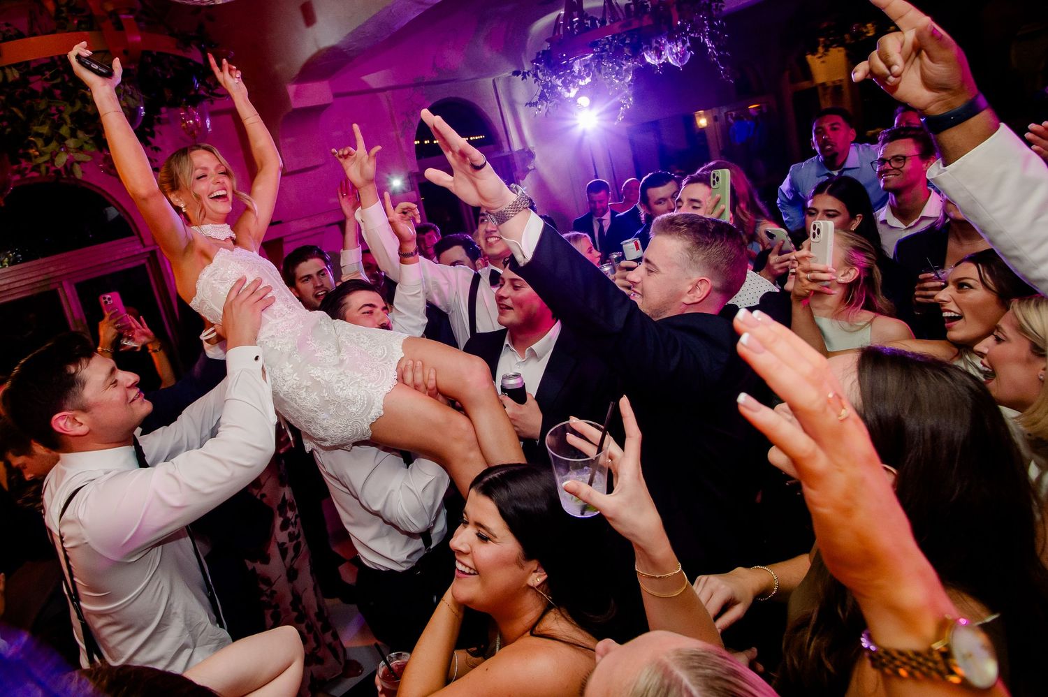 Energetic crowd dancing and celebrating at a nightclub with purple lighting and raised hands.
