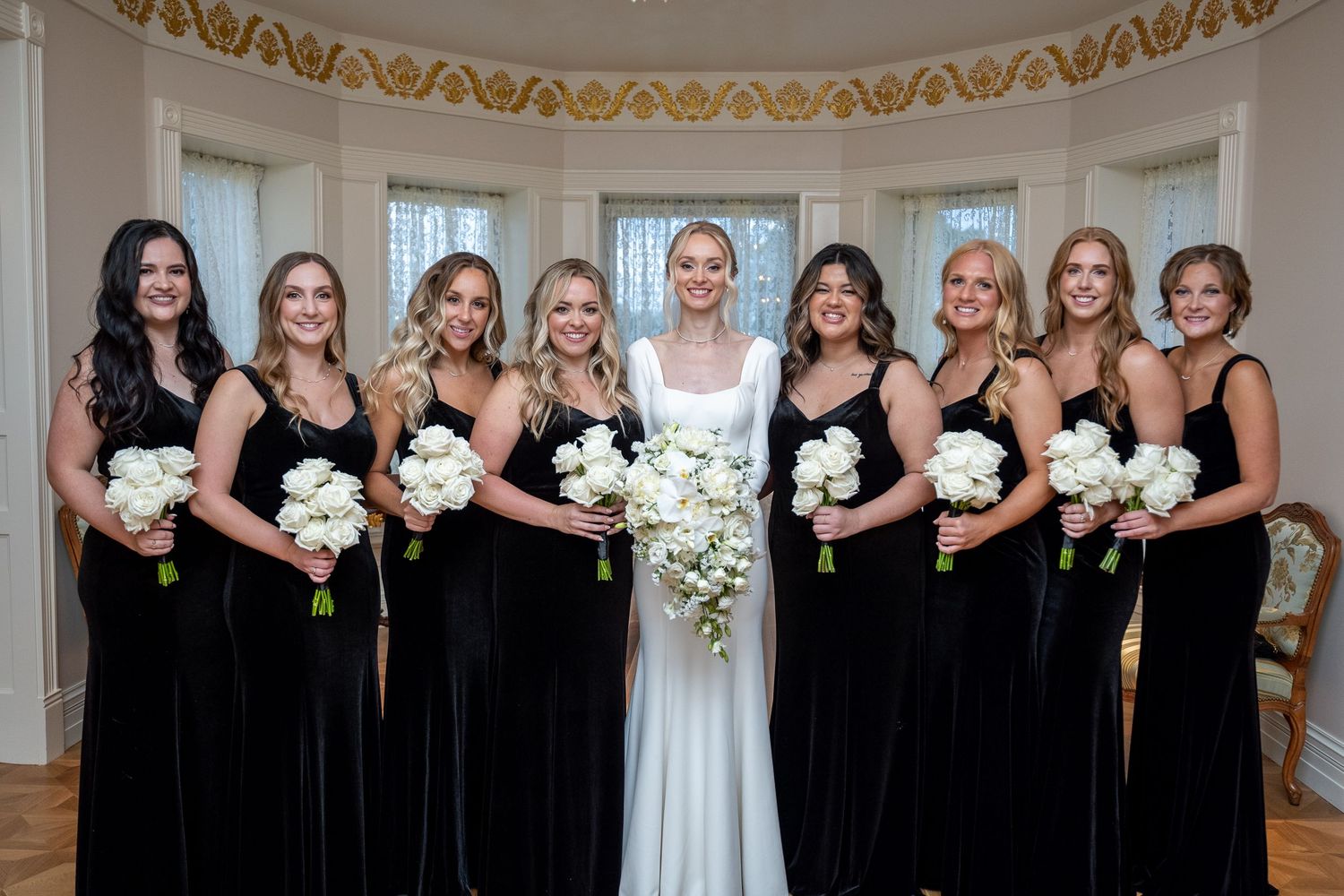 Wedding party in black bridesmaid dresses and white bridal gown pose together in an elegant room with ornate details.