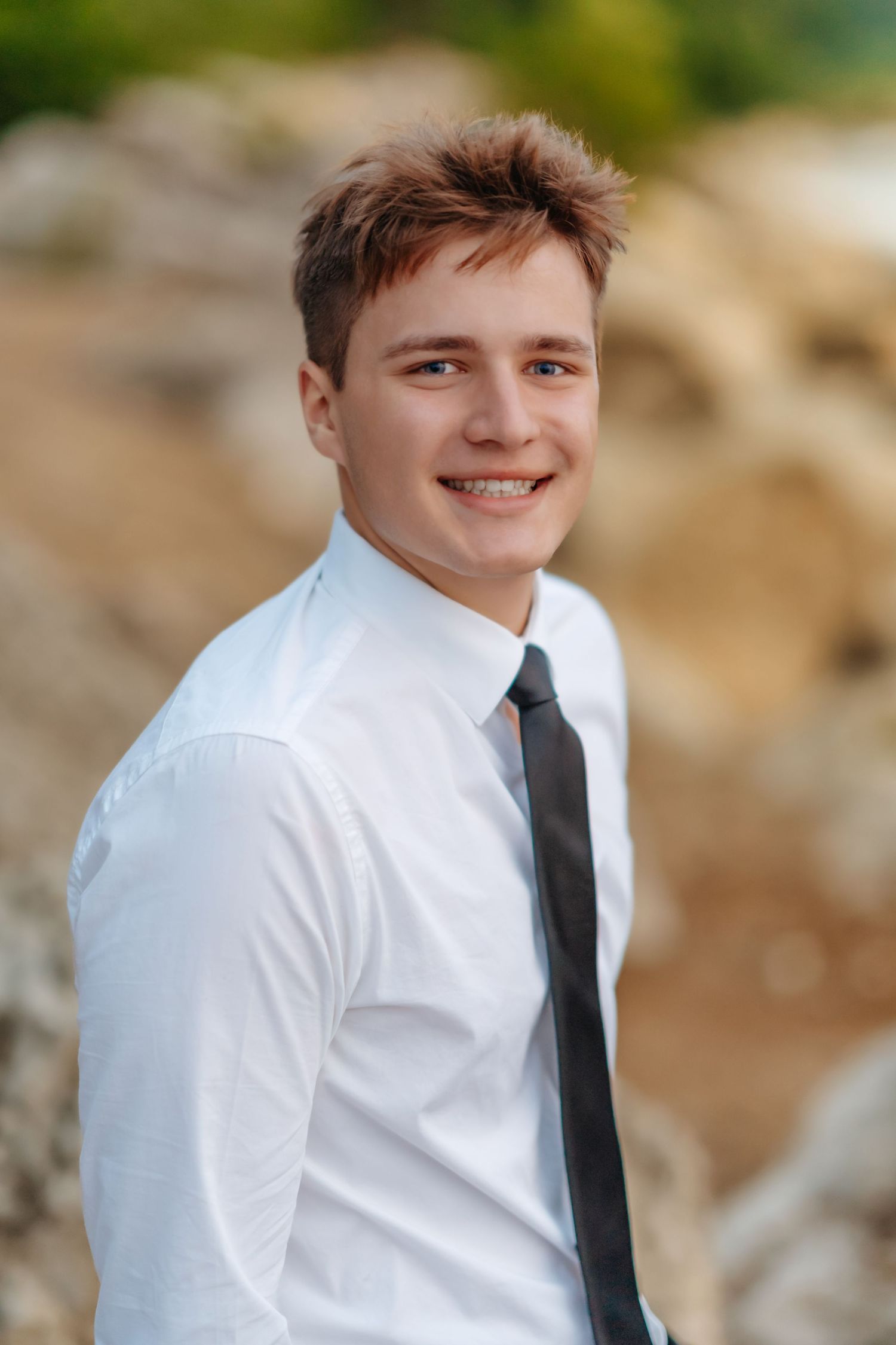 A person in a white dress shirt and black tie poses outdoors against a blurred natural background.