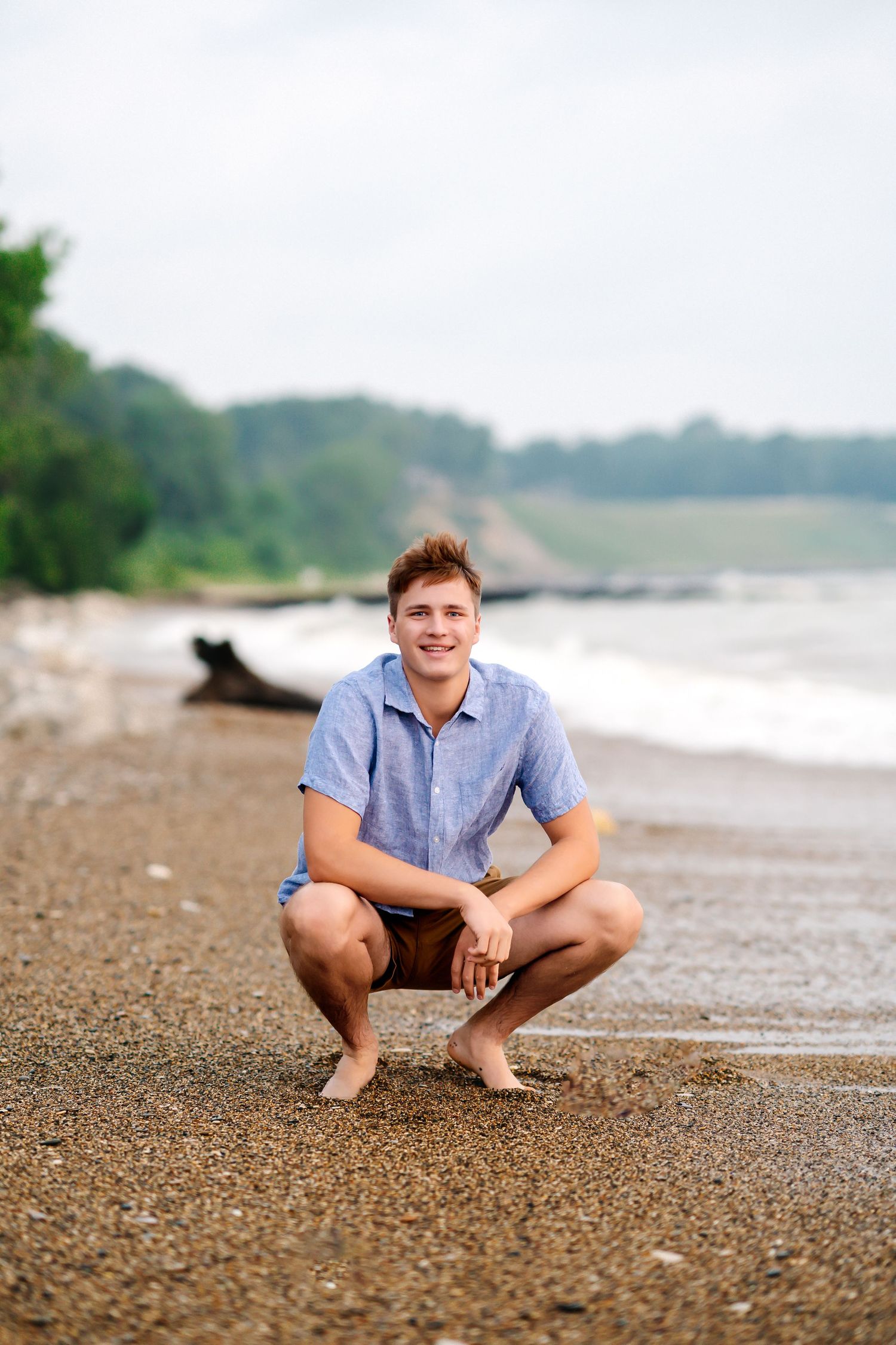 A person in a blue shirt sits on a pebbly beach near the water's edge on an overcast day.