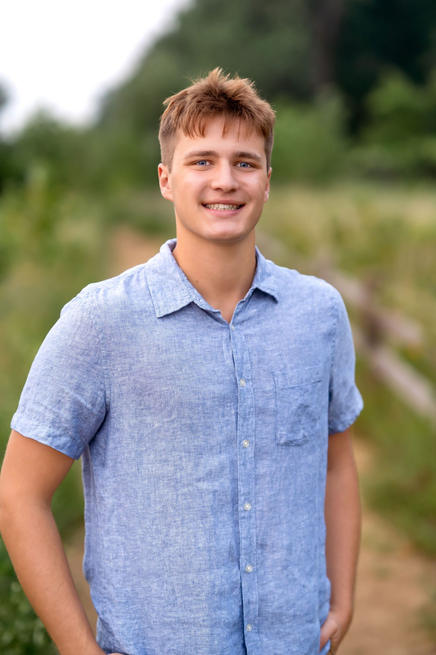 Person wearing a light blue chambray shirt poses outdoors on a sunny day with greenery in the background.