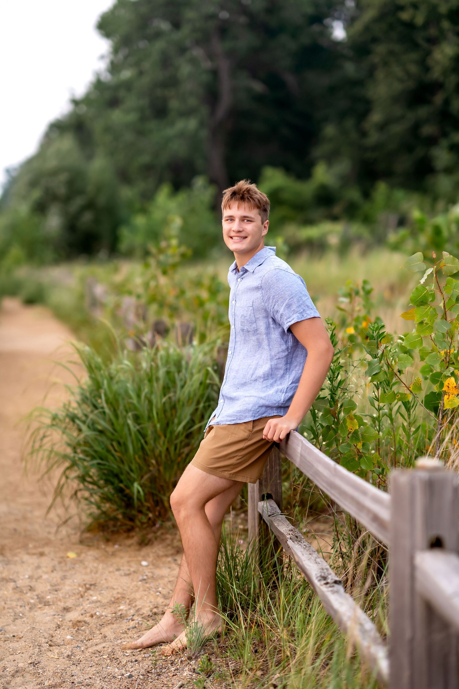 A person in a blue shirt and khaki shorts sits on a wooden railing surrounded by wildflowers and tall grass.