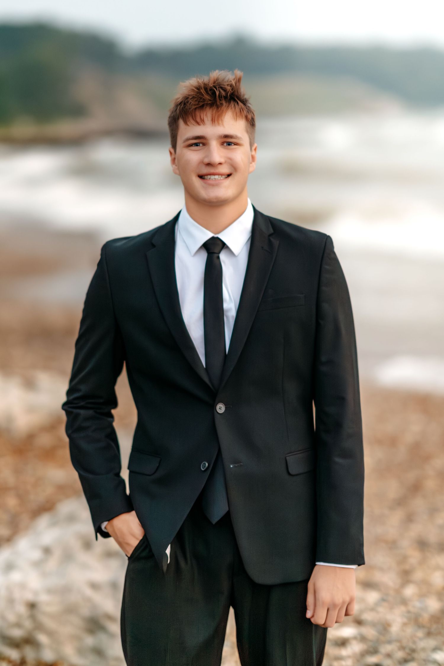 A person wearing a formal black suit and tie poses for photos on a beach with waves in the background.