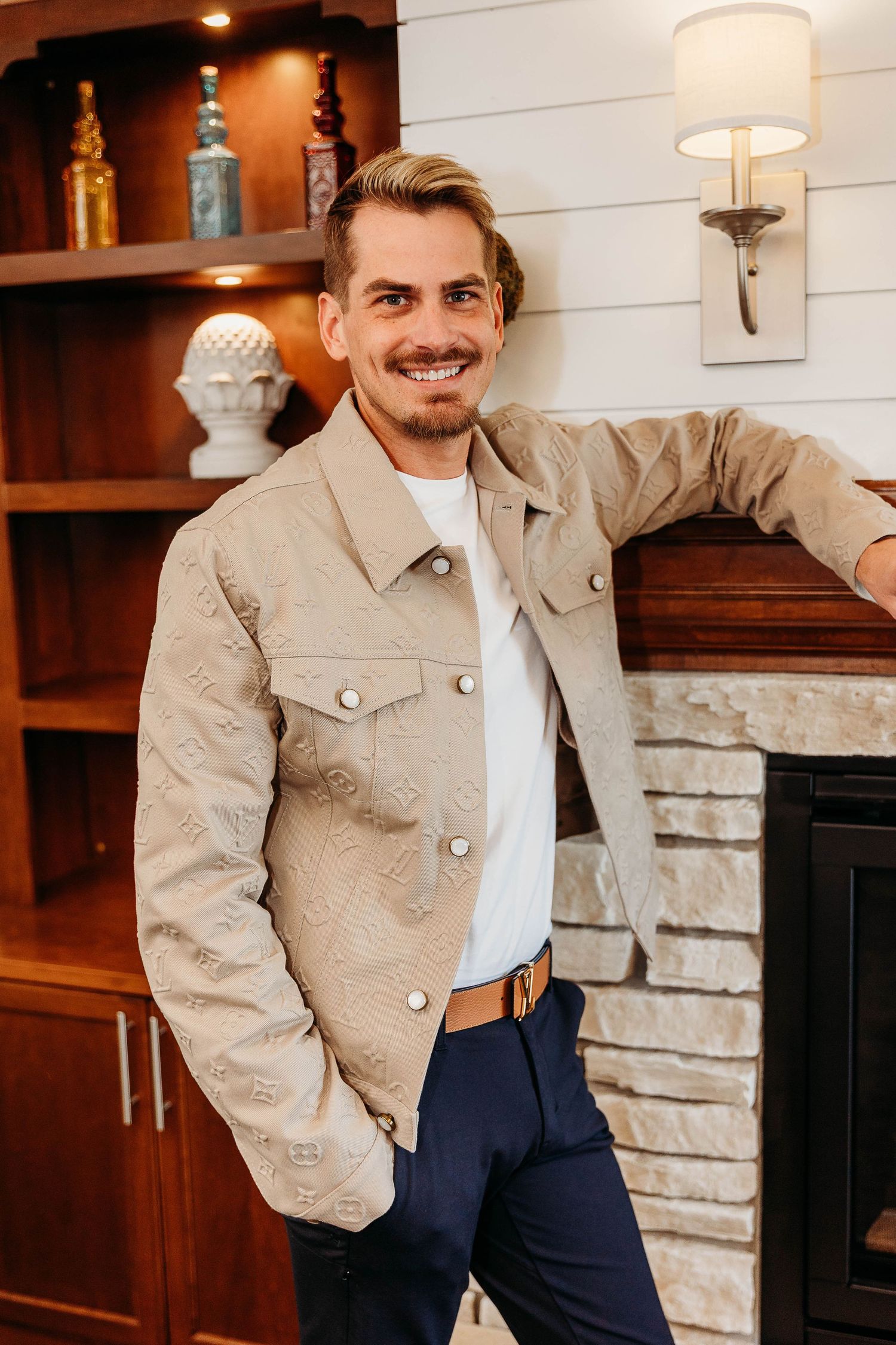 Stylish casual outfit featuring khaki jacket, white shirt and navy pants in modern home interior with wooden shelving.