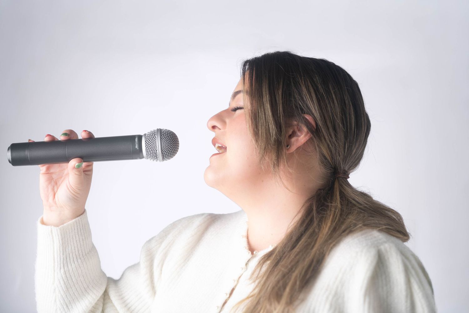Young woman singing passionately into microphone wearing white sweater on light background.
