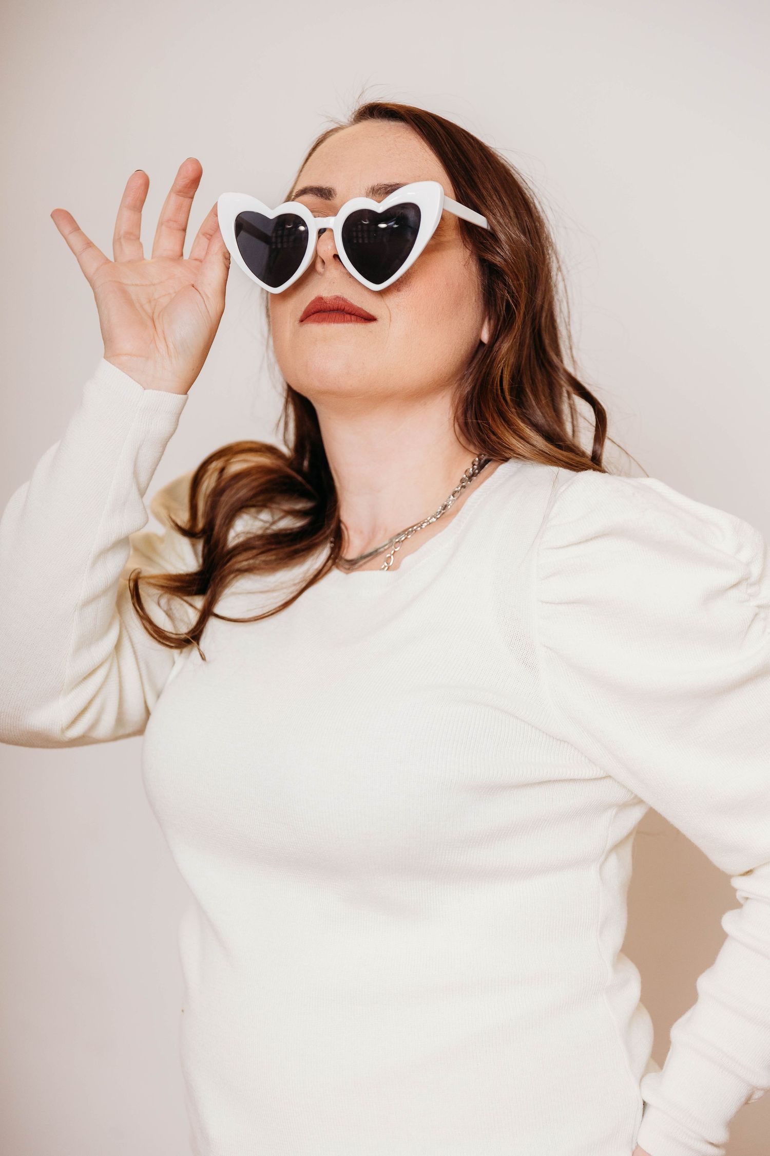 A person in a white sweater strikes playful poses against a light background while wearing heart-shaped sunglasses.