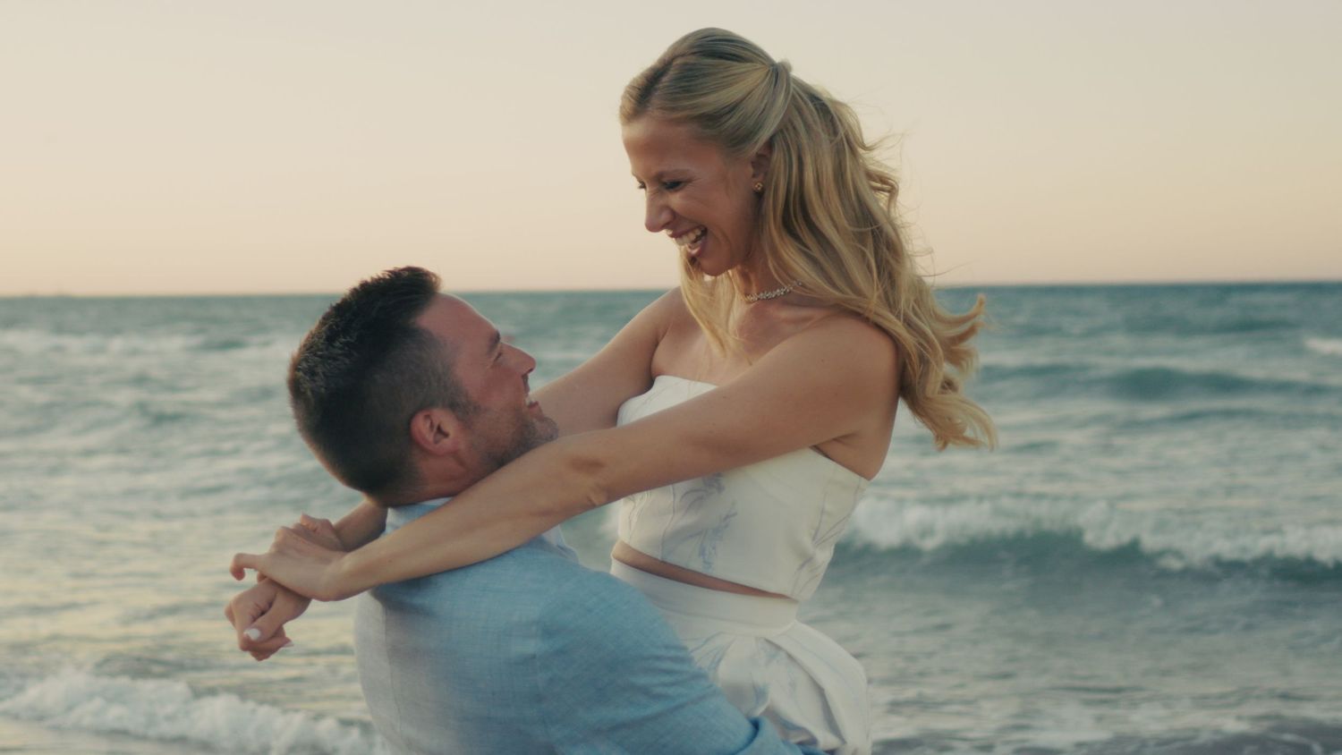 A joyful couple embraces and spins on a sandy beach at sunset with ocean waves in the background.